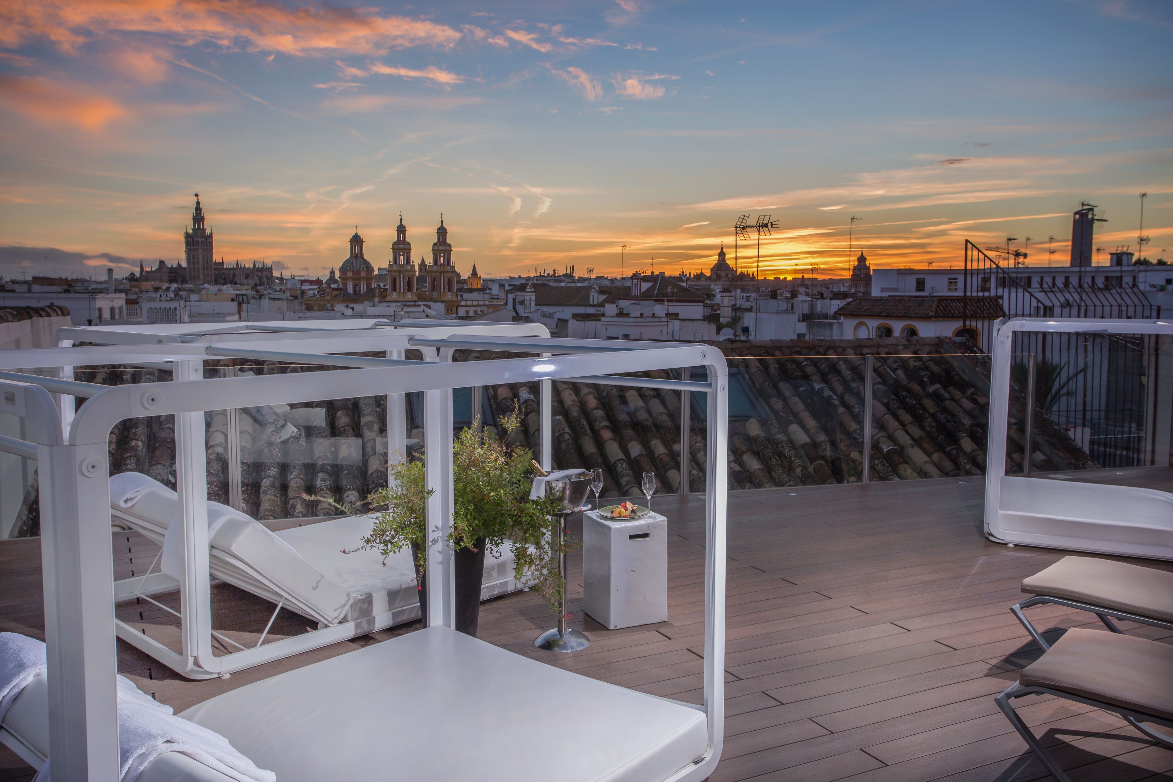A rooftop lounge with sun loungers, a small table with a plant and wine glass, and a view of the city skyline at sunset.