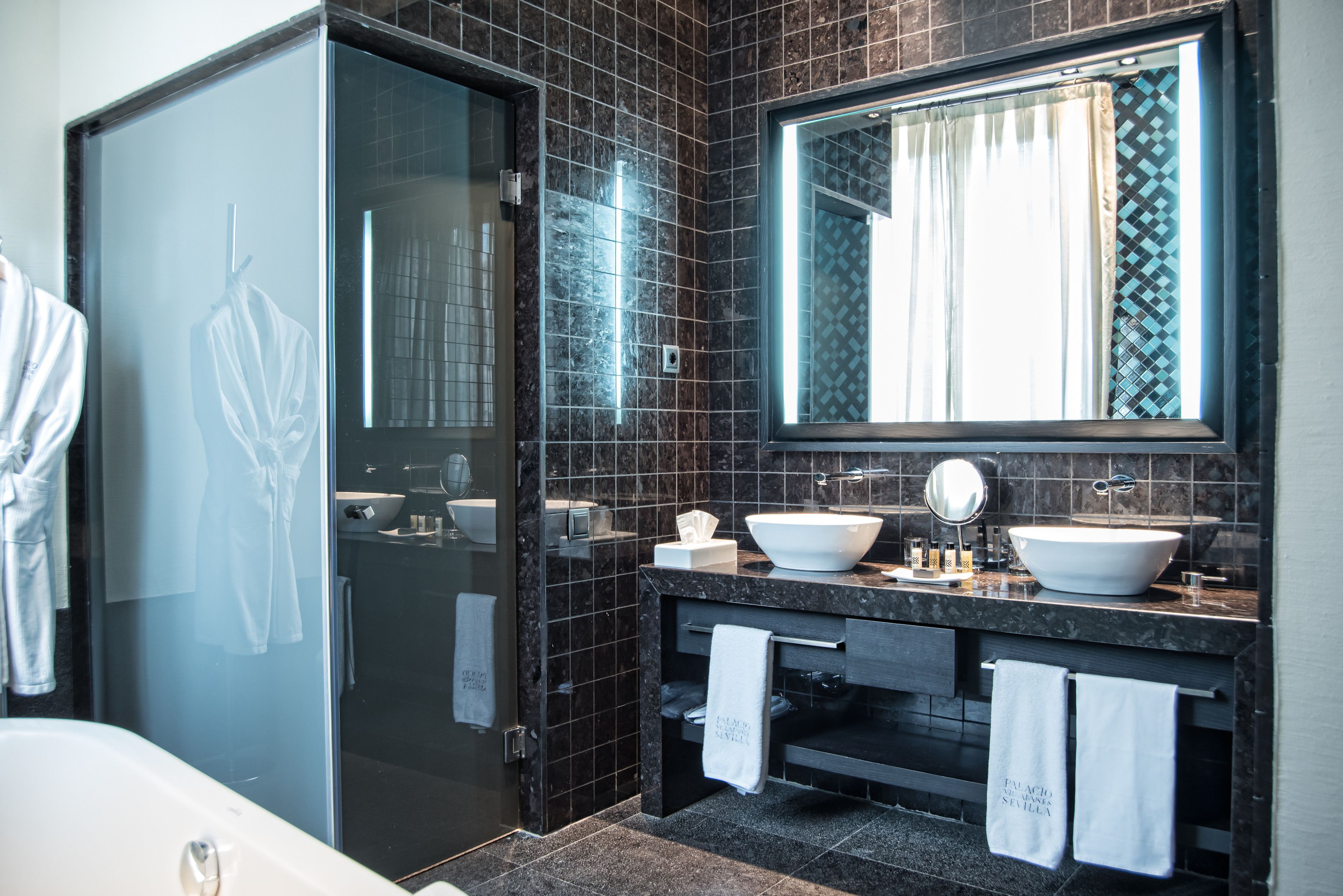 A modern bathroom with a glass shower enclosure, two sinks, a large mirror, and dark tiled walls.