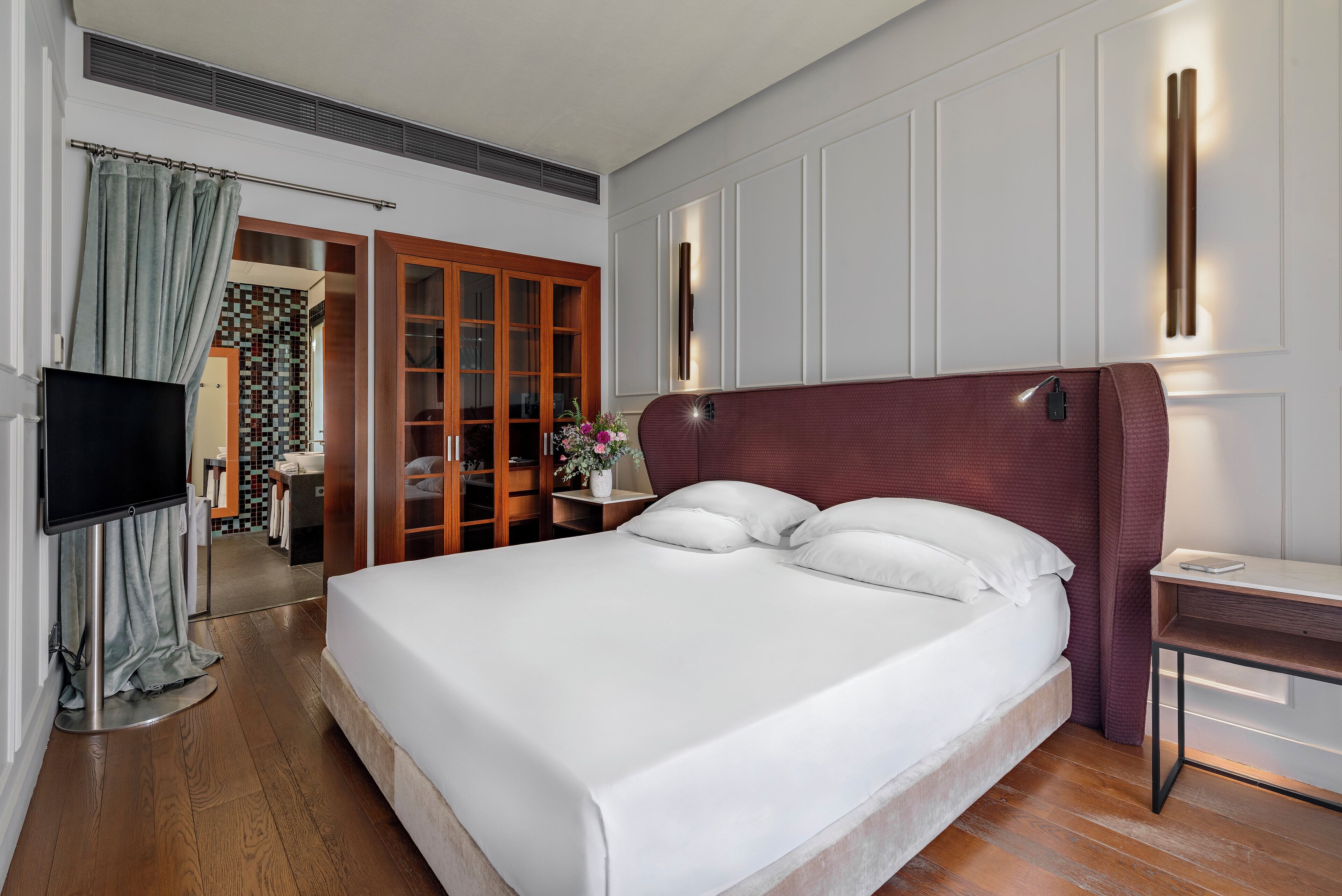 A modern hotel room with a large bed, a TV, a desk, and a bathroom visible through an open door.