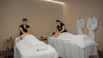 A spa room with two massage tables, each covered with a white towel and a logo. There are two towels hanging on the wall and a small table with bottles and a bowl.