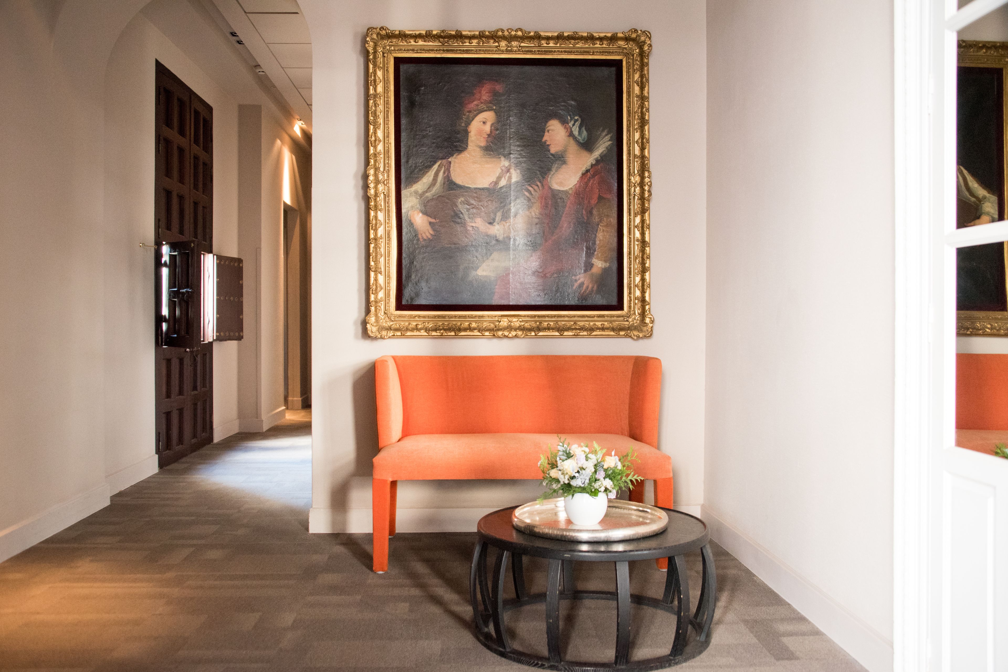 A hallway with a sofa, a round table with a vase, and a framed painting on the wall.