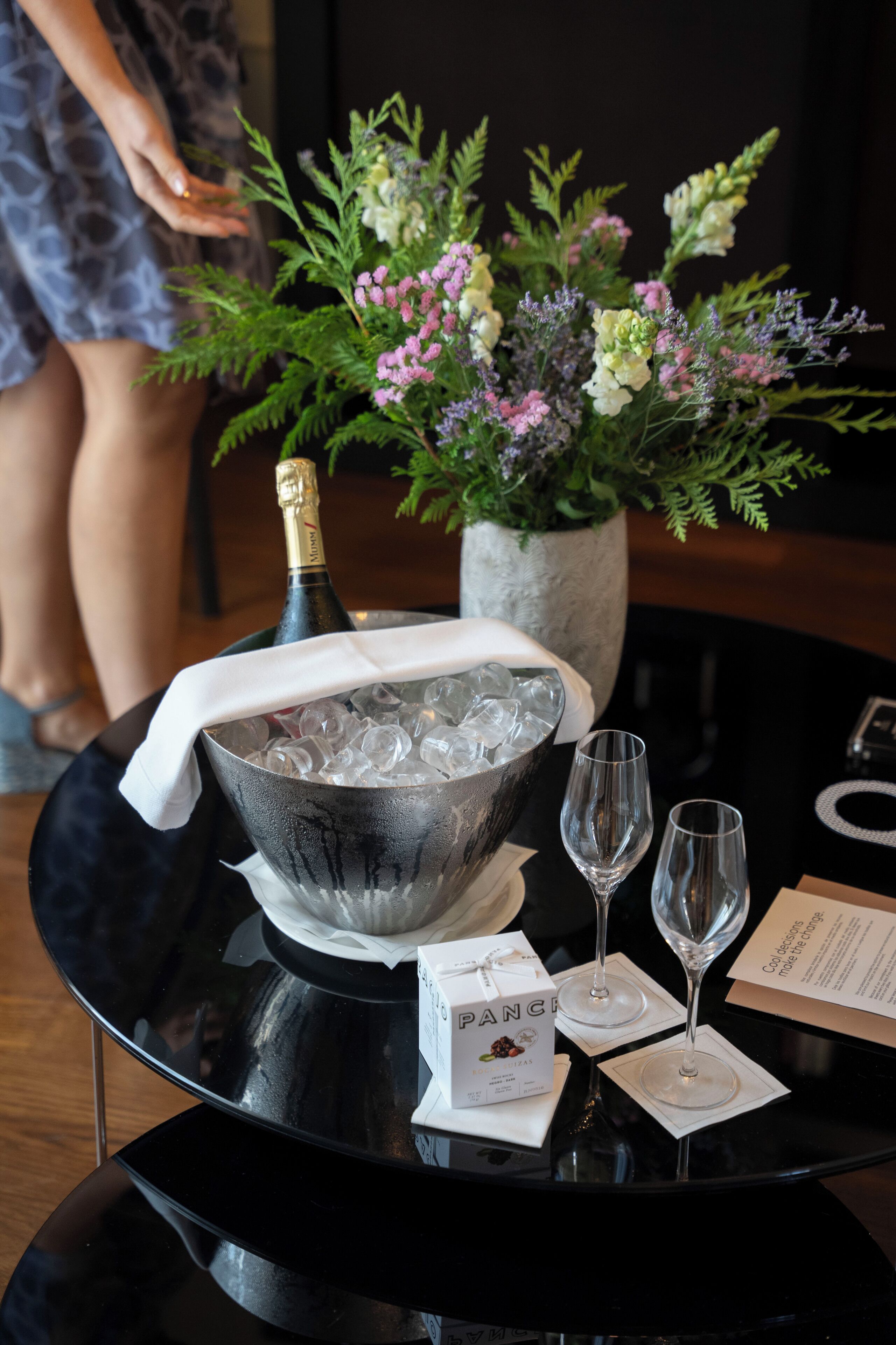 Ein Tisch mit einer Champagnerflasche, Eiswürfeln, zwei Gläsern, einem Blumenstrauß und einer mit ‚PANG‘ beschrifteten Schachtel.