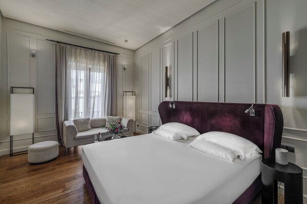 Habitación de hotel con una cama grande, un sofá, una mesita con un florero y una ventana con cortinas.