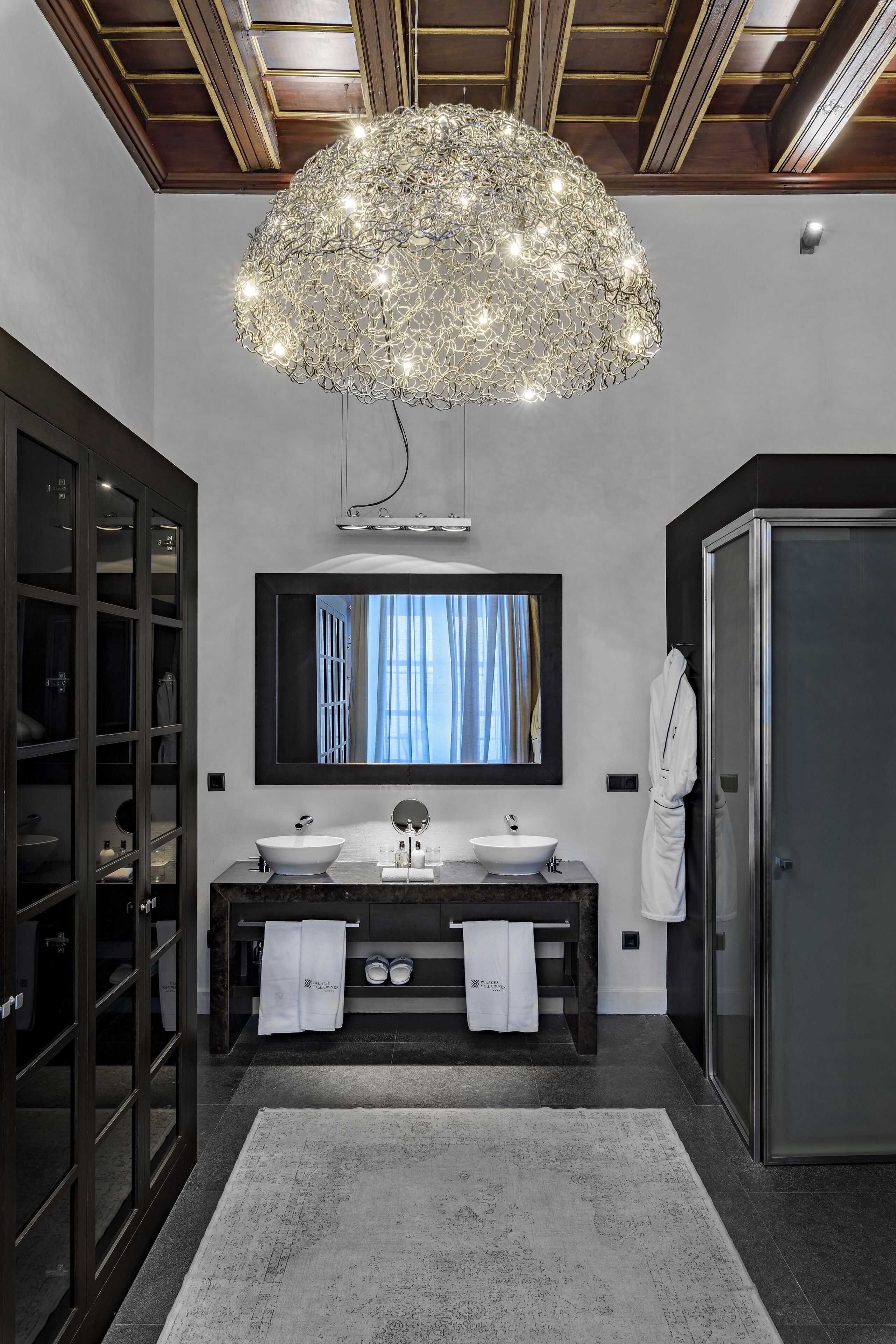 A modern bathroom with a large chandelier, dual sinks, and a mirror above the vanity.