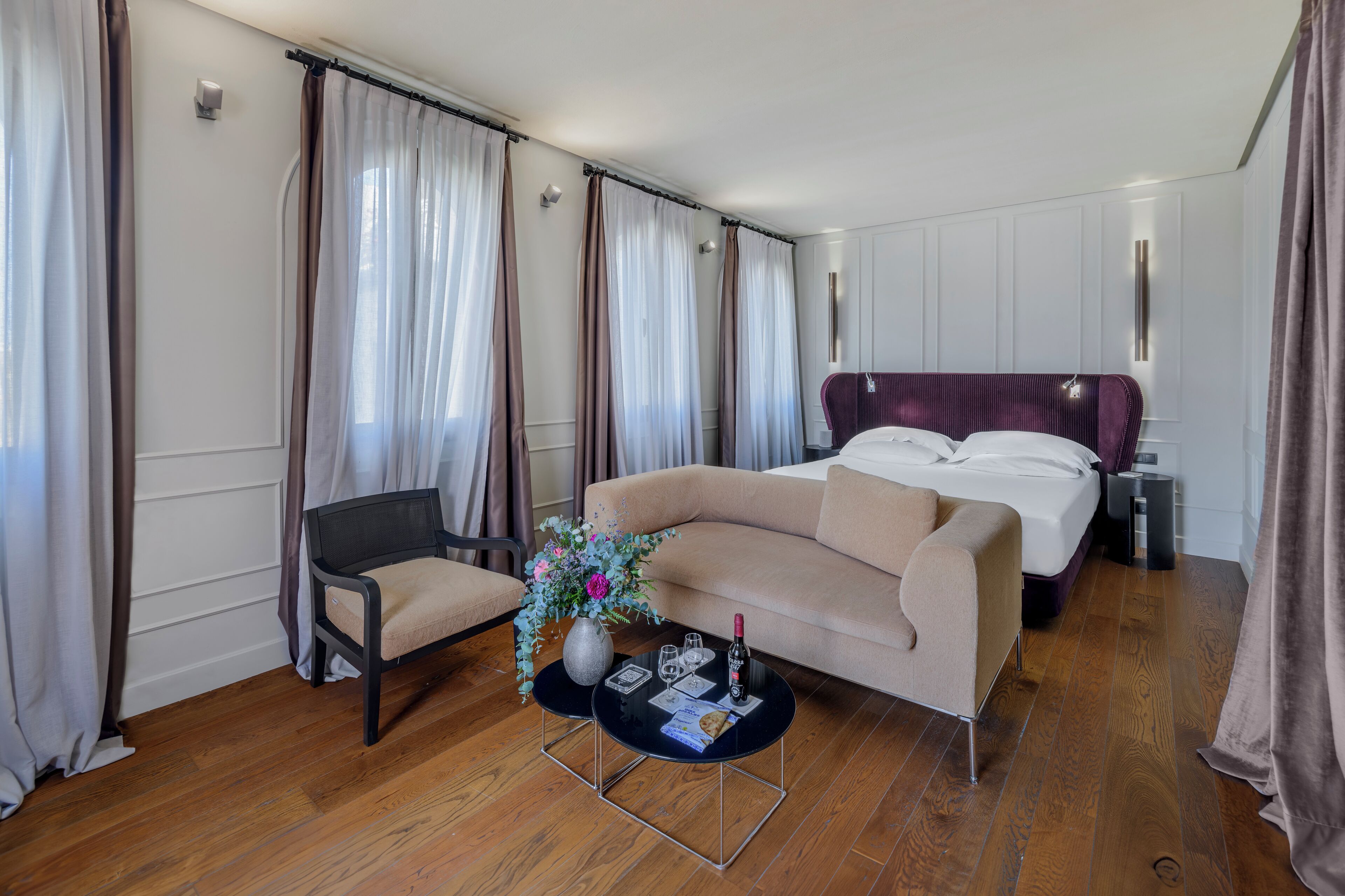 A hotel room with a sofa, a chair, a small table with a vase and a bottle, and a bed with a headboard.