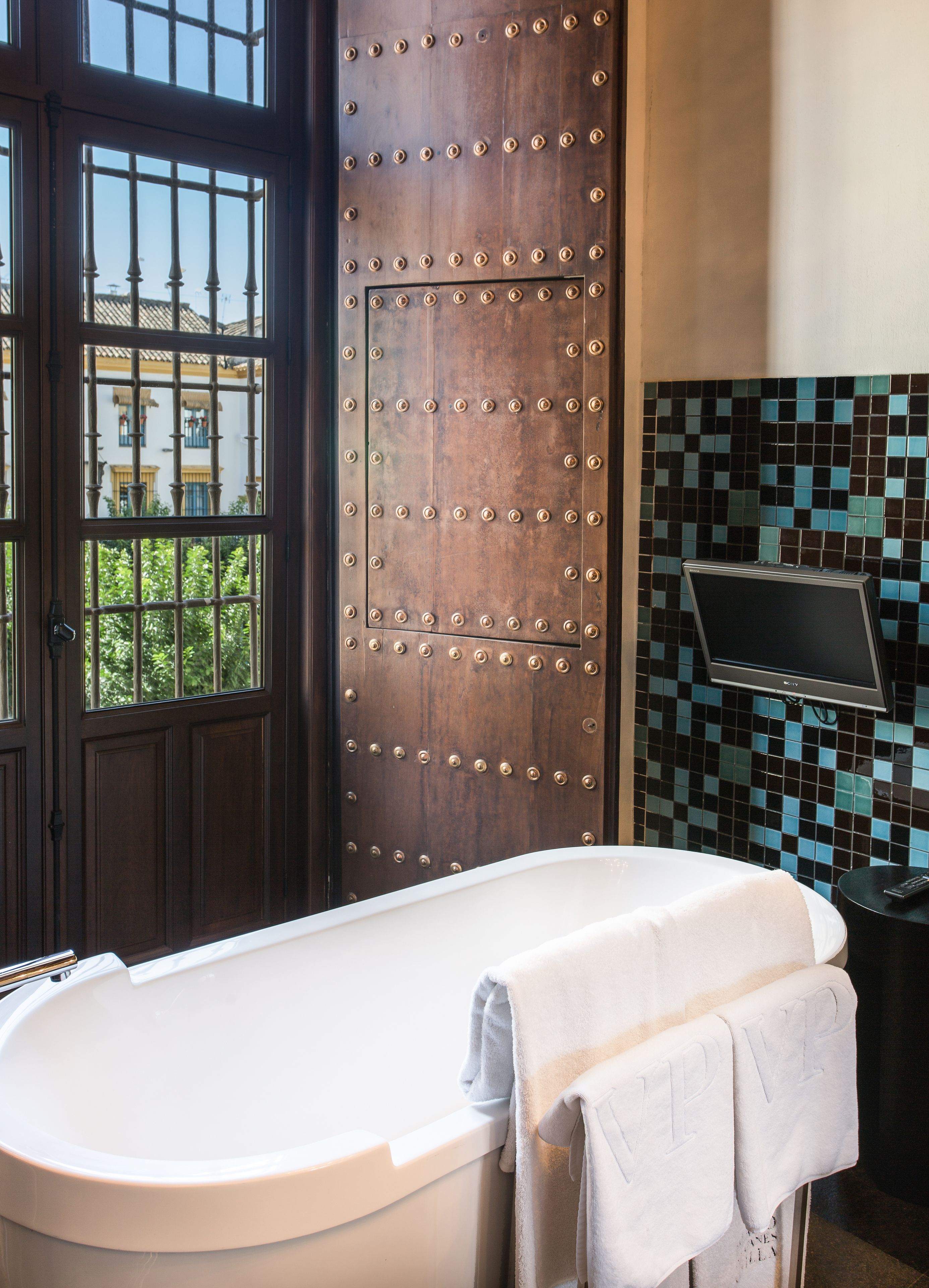 A bathroom with a bathtub, a wooden door with metal studs, and a tiled wall with a television.