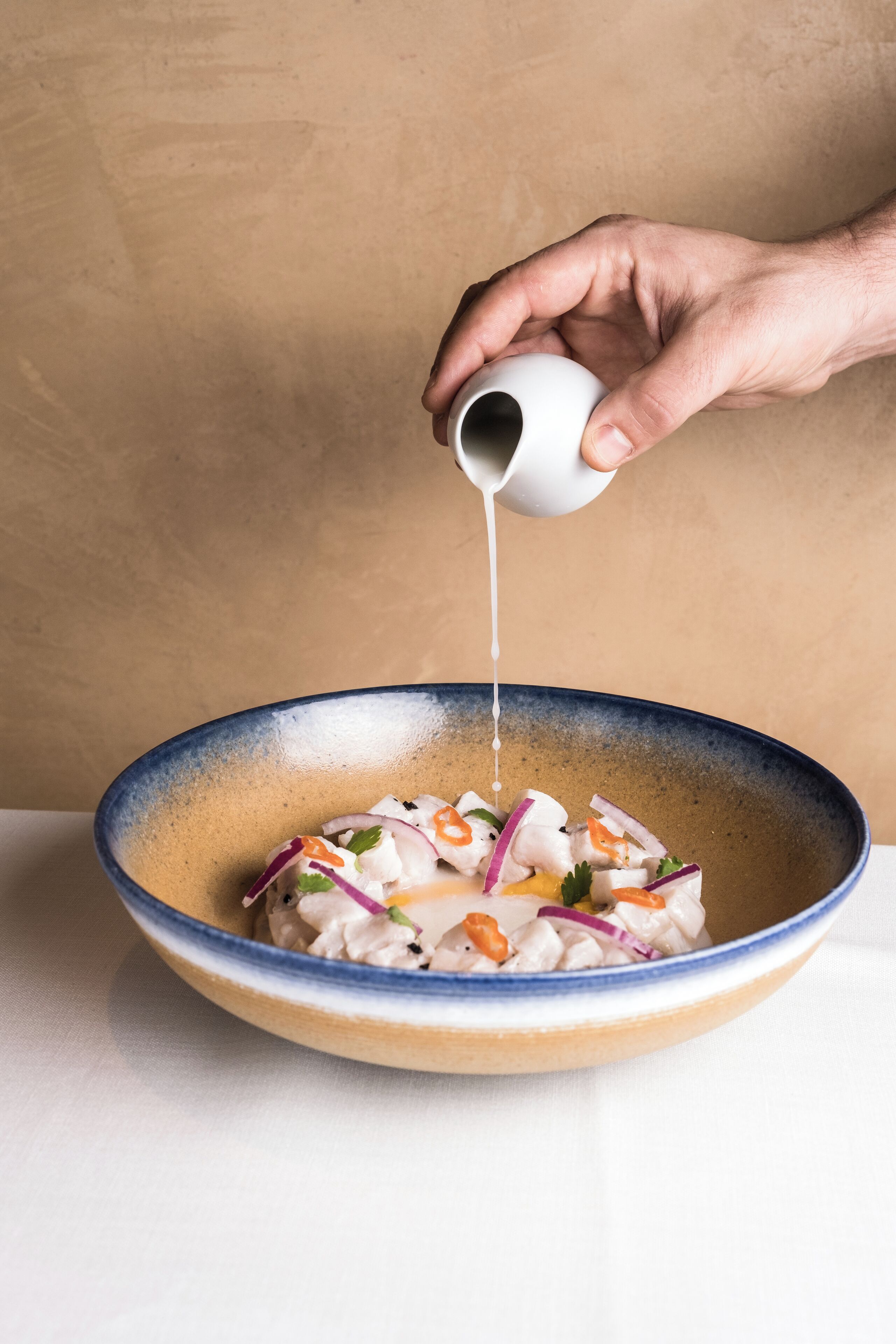 A hand pouring a white liquid into a bowl containing a seafood salad with vegetables.