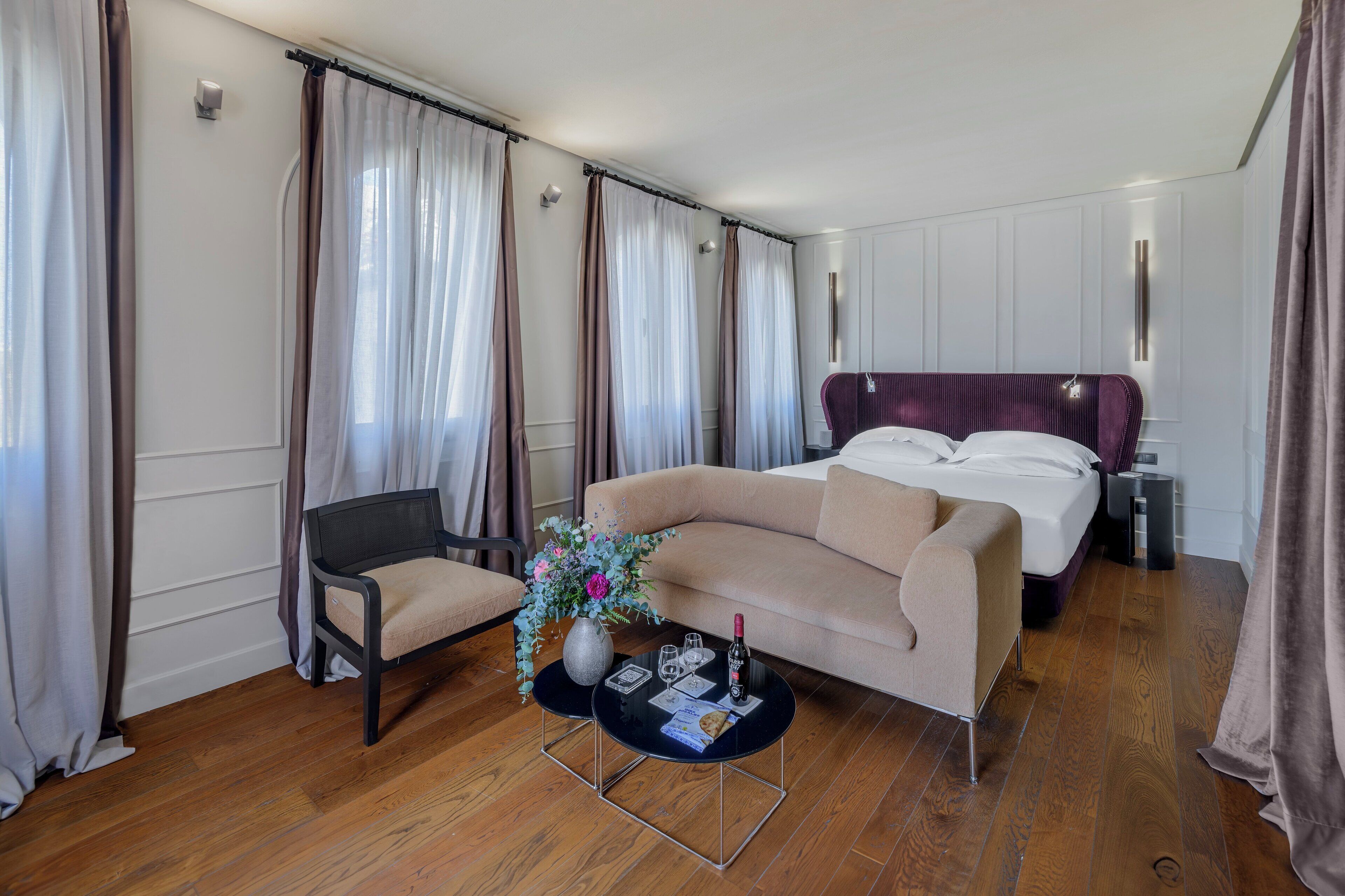 A hotel room with a bed, a sofa, a chair, a coffee table with a vase of flowers, and a wine bottle.