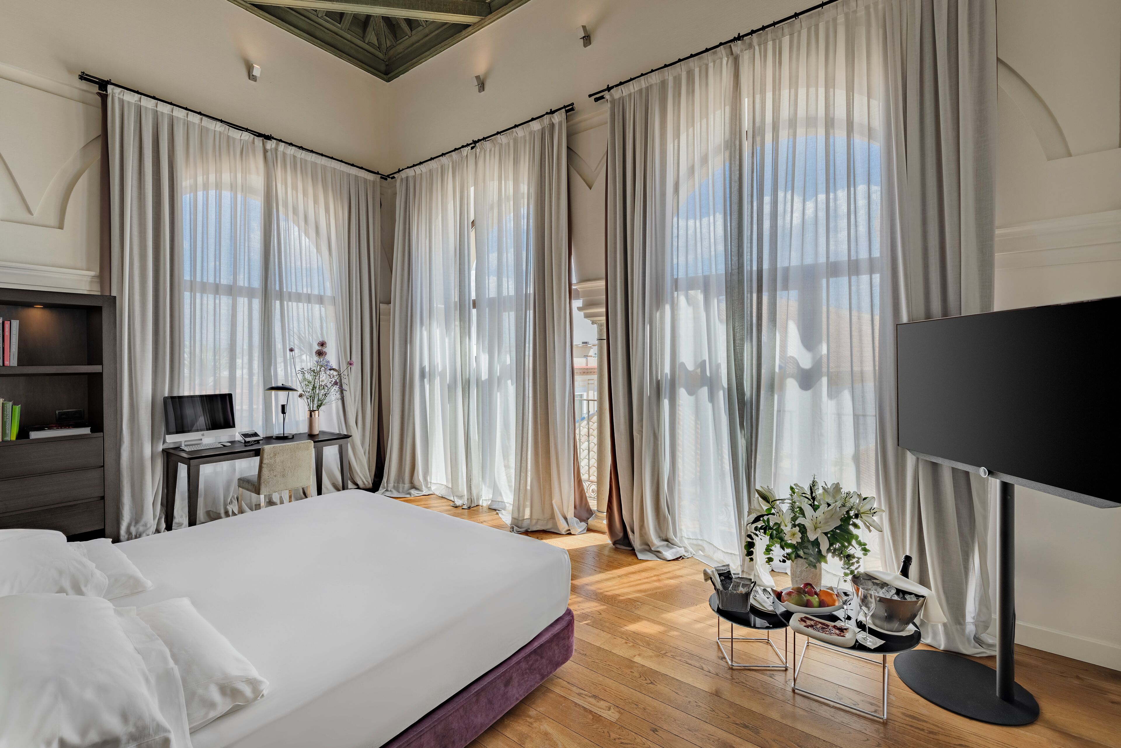 A modern hotel room with a large bed, a desk with a computer, a flat-screen TV, and a dining area with a table set for two.