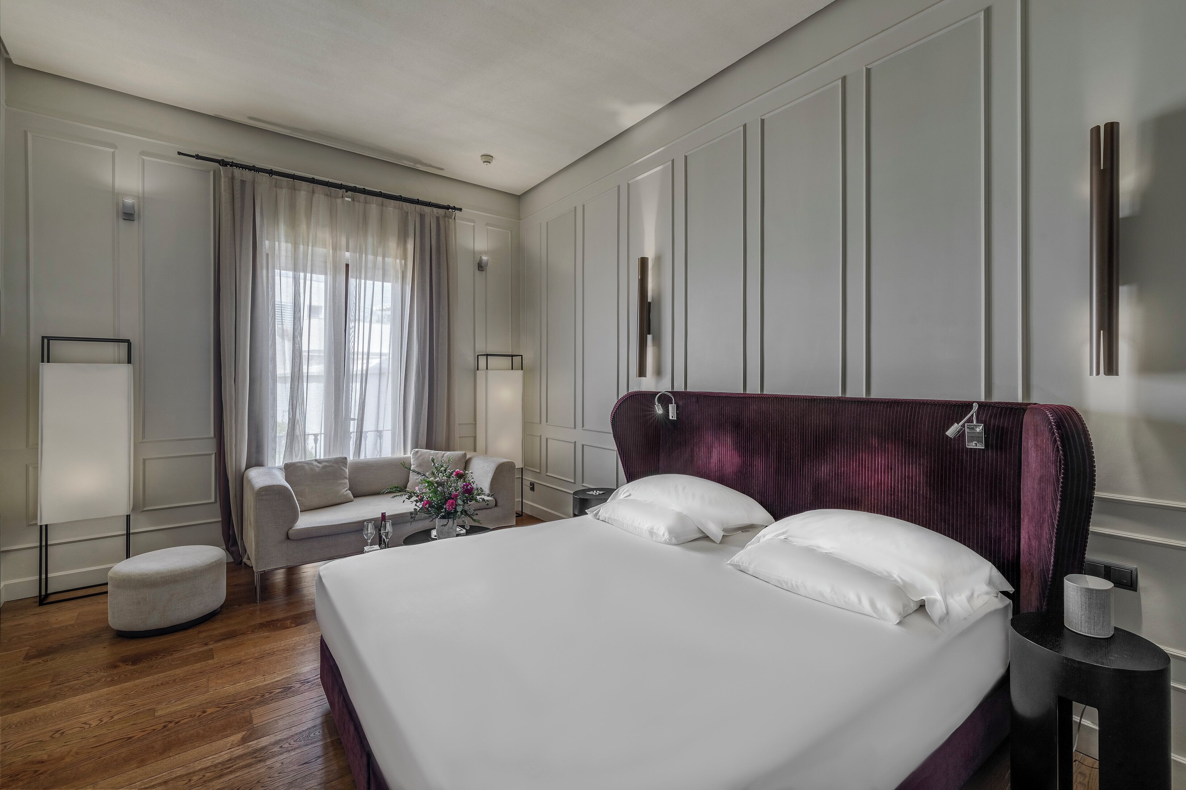 A hotel room with a large bed, a sofa, a small table with a vase of flowers, and a window with curtains.