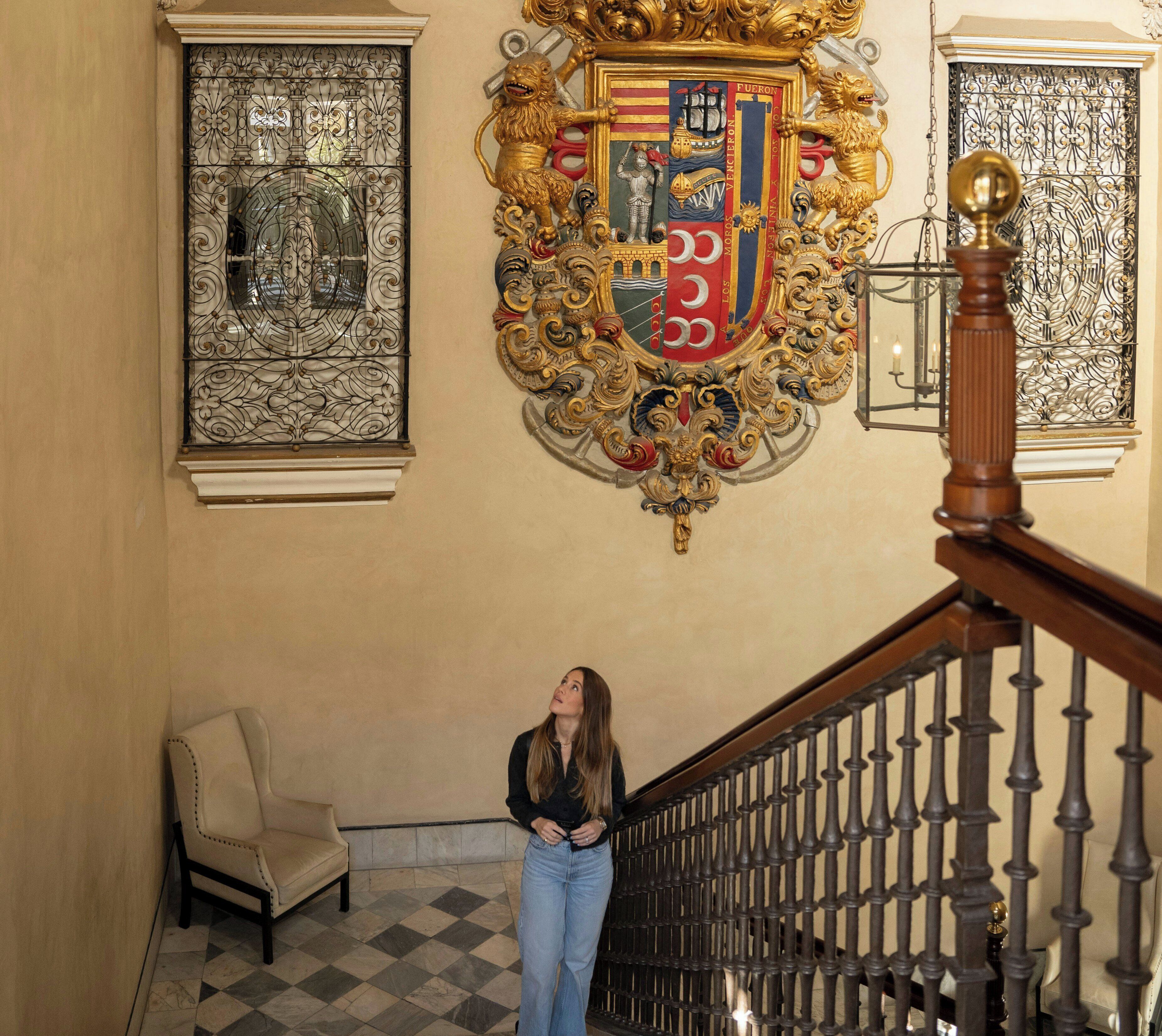 A woman standing on a staircase with a decorative coat of arms above her.