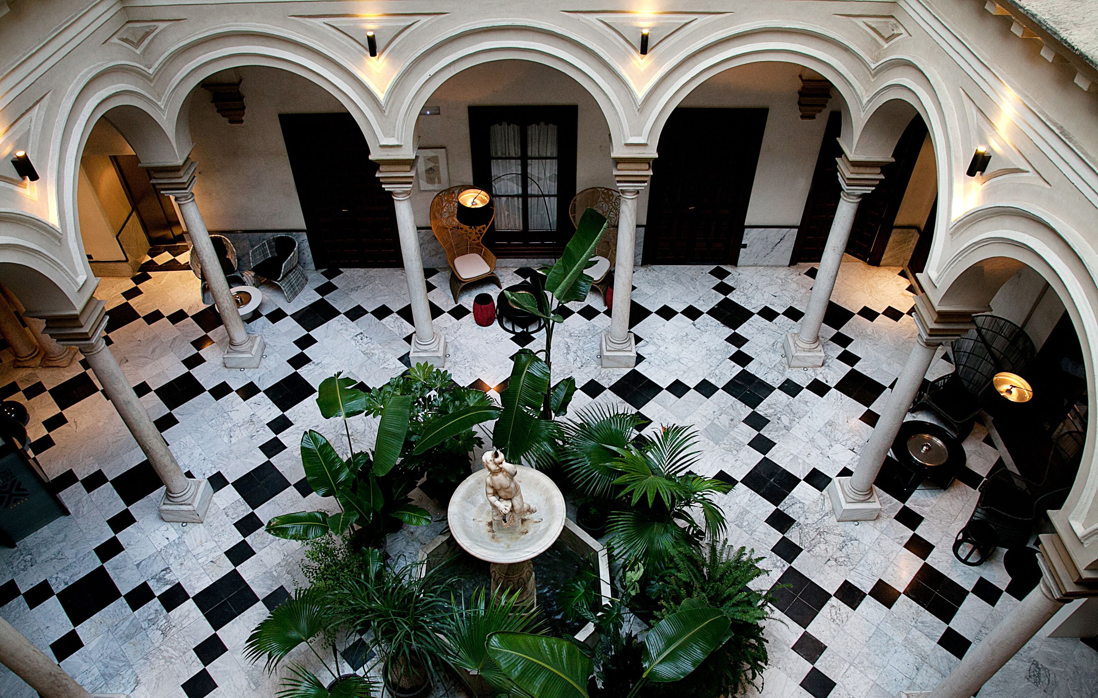 Un patio con suelo a cuadros, columnas y una fuente central rodeada de follaje.