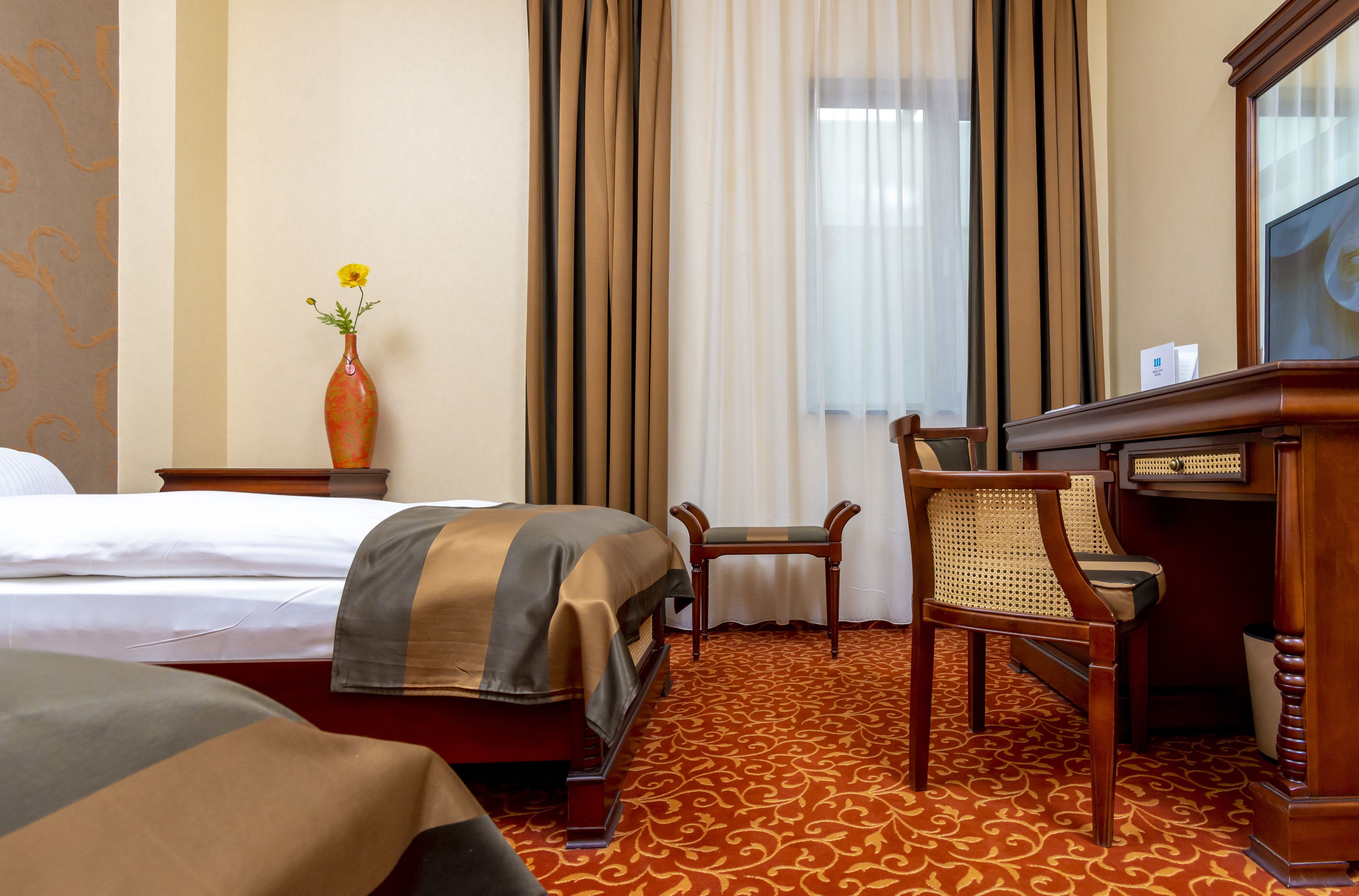 A hotel room with a bed, a desk, a chair, a vase with a flower, and a window with curtains.