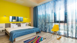 A modern hotel room with a large bed, a vibrant yellow accent wall, a city view through sheer curtains, and a colorful striped rug. - West City Hotel (Floresti)