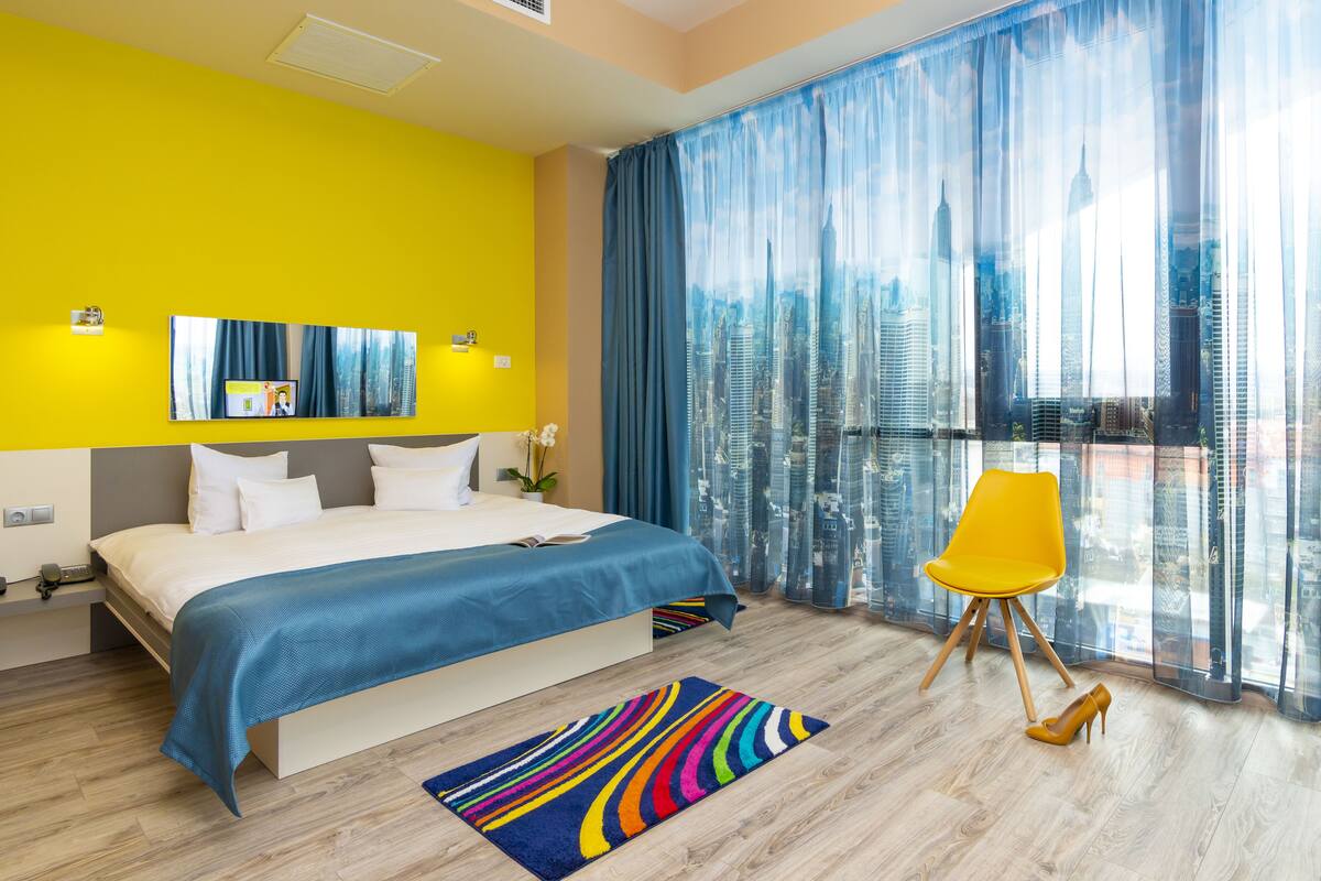 A modern hotel room with a large bed, a vibrant yellow accent wall, a city view through sheer curtains, and a colorful striped rug.