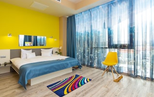 A modern hotel room with a large bed, a vibrant yellow accent wall, a city view through sheer curtains, and a colorful striped rug.