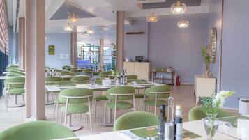 A restaurant with green chairs and tables, pendant lighting, and a view of the city through large windows.