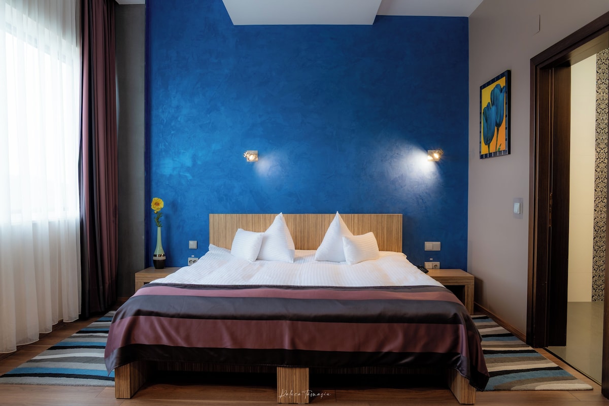 A hotel room with a large bed, a nightstand, a vase with a flower, and a painting on the wall.