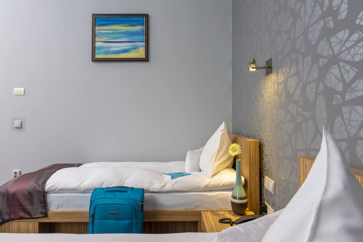 A hotel room with two beds, a blue suitcase, a vase with a yellow flower, and a painting on the wall.