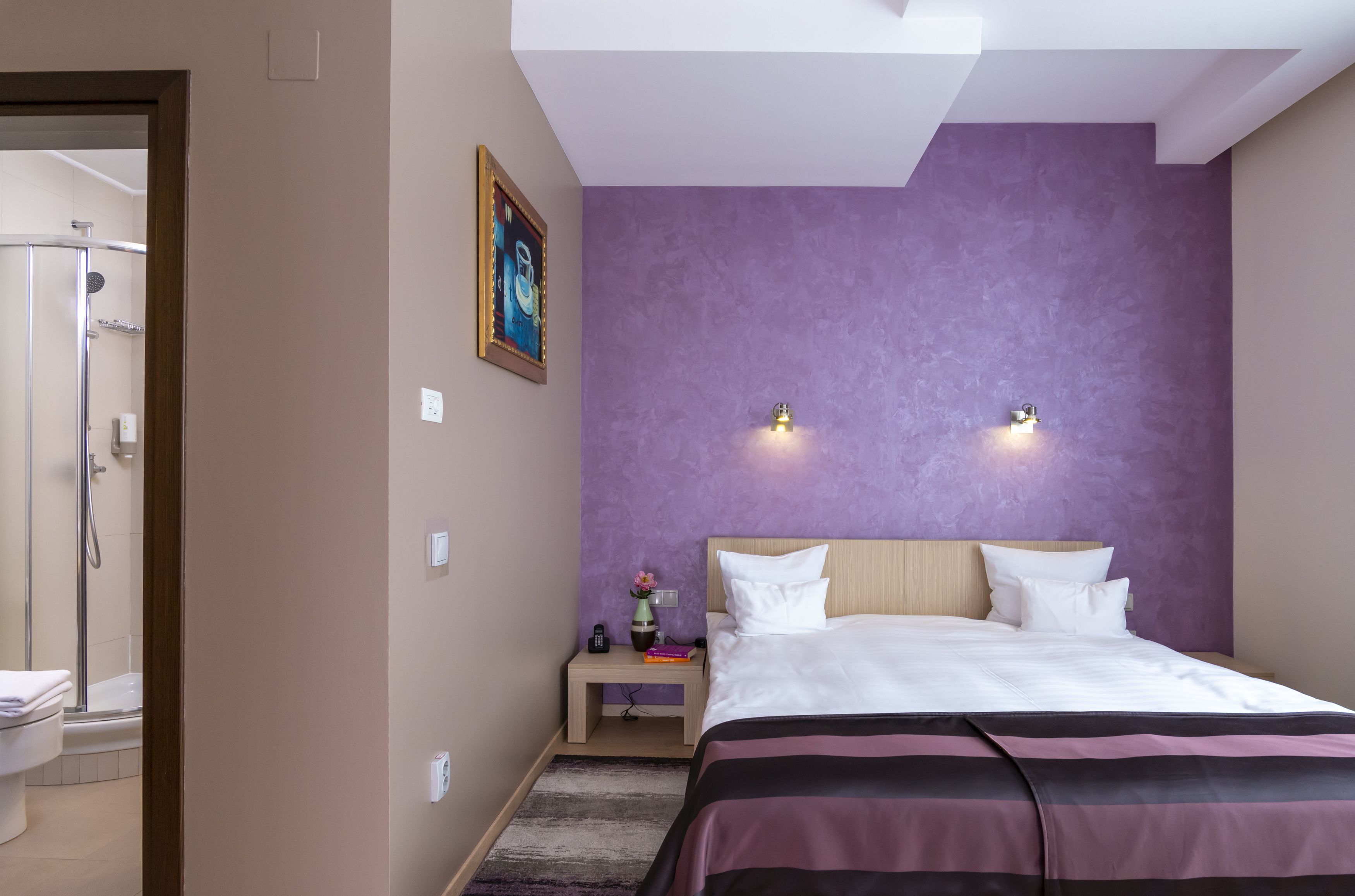 Habitación de hotel con cama, mesita de noche con un florero y libros, ducha y una pared de acento color púrpura.