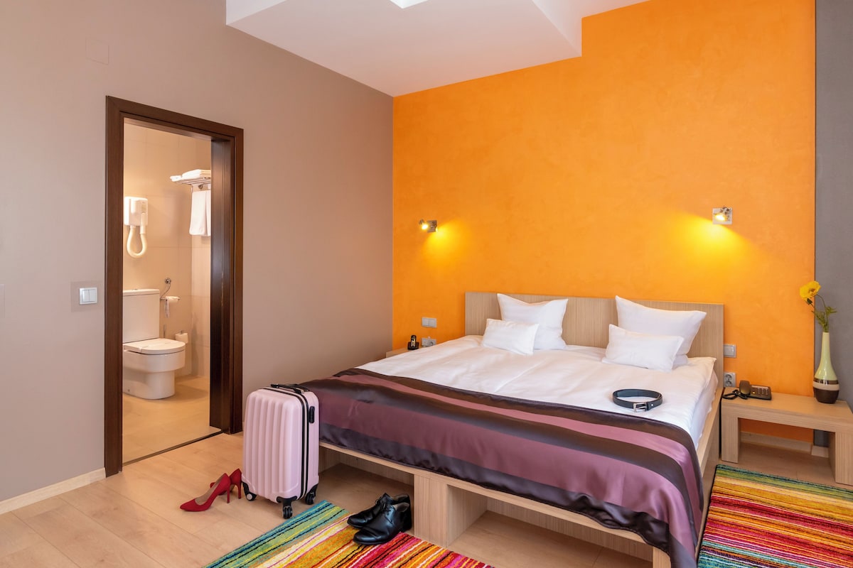 A hotel room with a bed, a suitcase, a pair of shoes, a bathroom with a toilet and shower, and a small table with a vase.