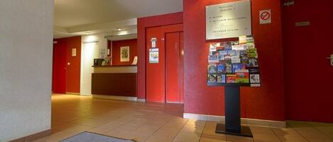 Zona de recepción de hotel con una pared roja, un mostrador y un tablón de anuncios con varios avisos.