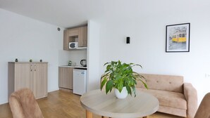 A modern living room with a round wooden table, two beige sofas, and a potted plant. A kitchen with wooden cabinets and a white refrigerator is visible in the background. - KOSY Appart’Hôtels – La Maison des Chercheurs (Vandœuvre-lès-Nancy)