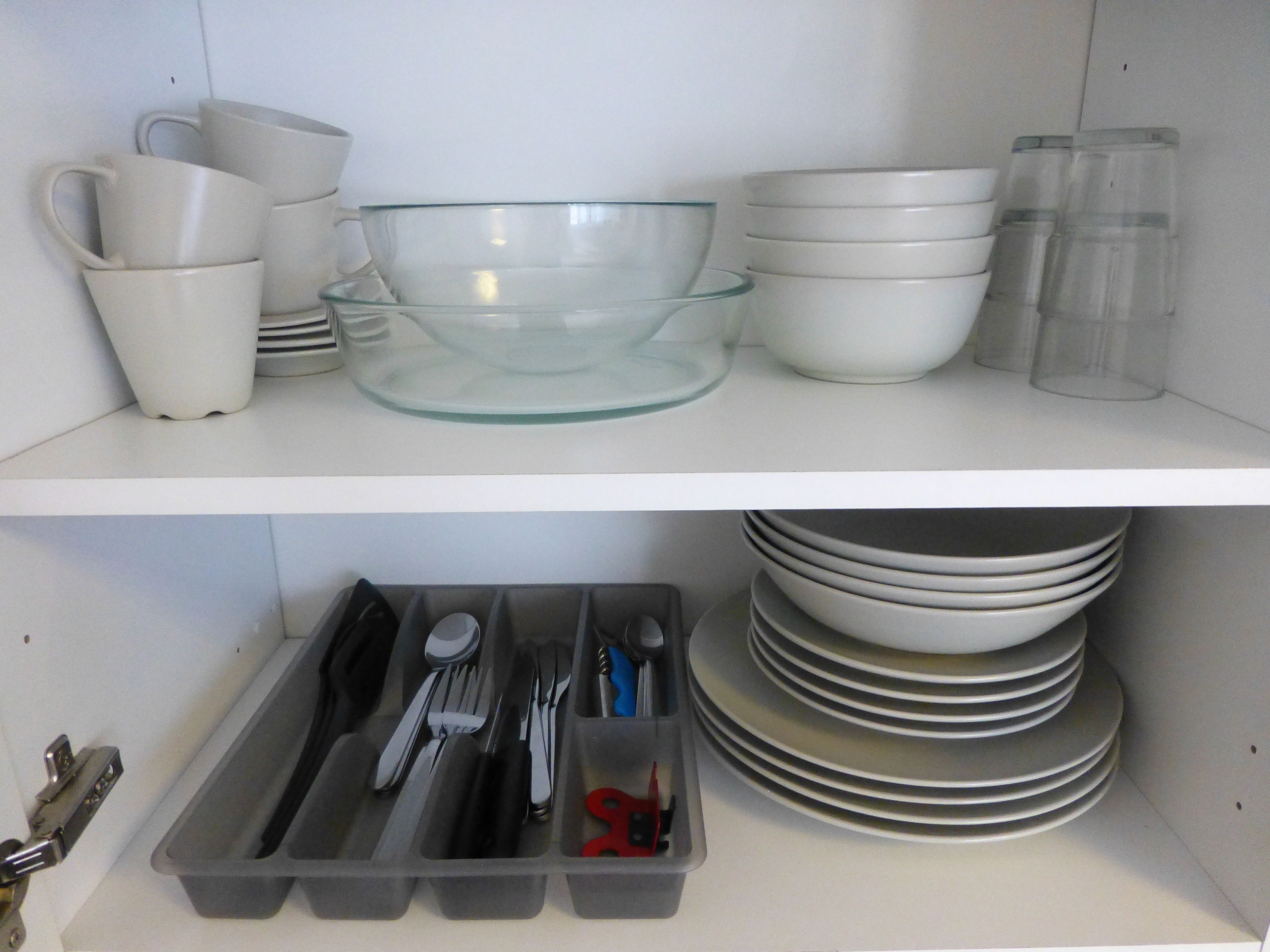 Una cucina con ripiani bianchi che contengono diverse ciotole, tazze e bicchieri. Sotto, un cassetto con utensili e piatti.