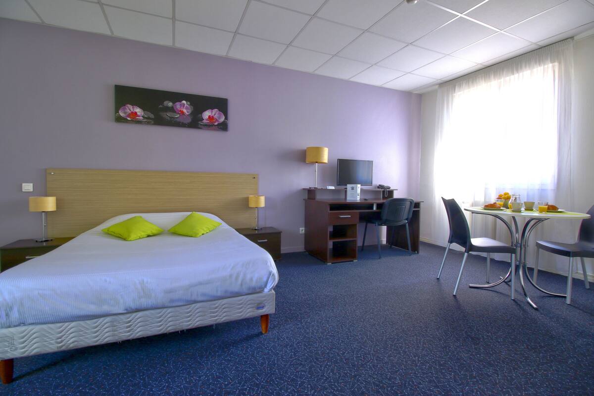 A hotel room with a bed, desk, chair, and a painting on the wall.