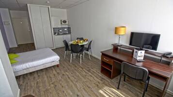 A hotel room with a bed, a desk, a TV, a lamp, a chair, a table with fruits, and a kitchenette.