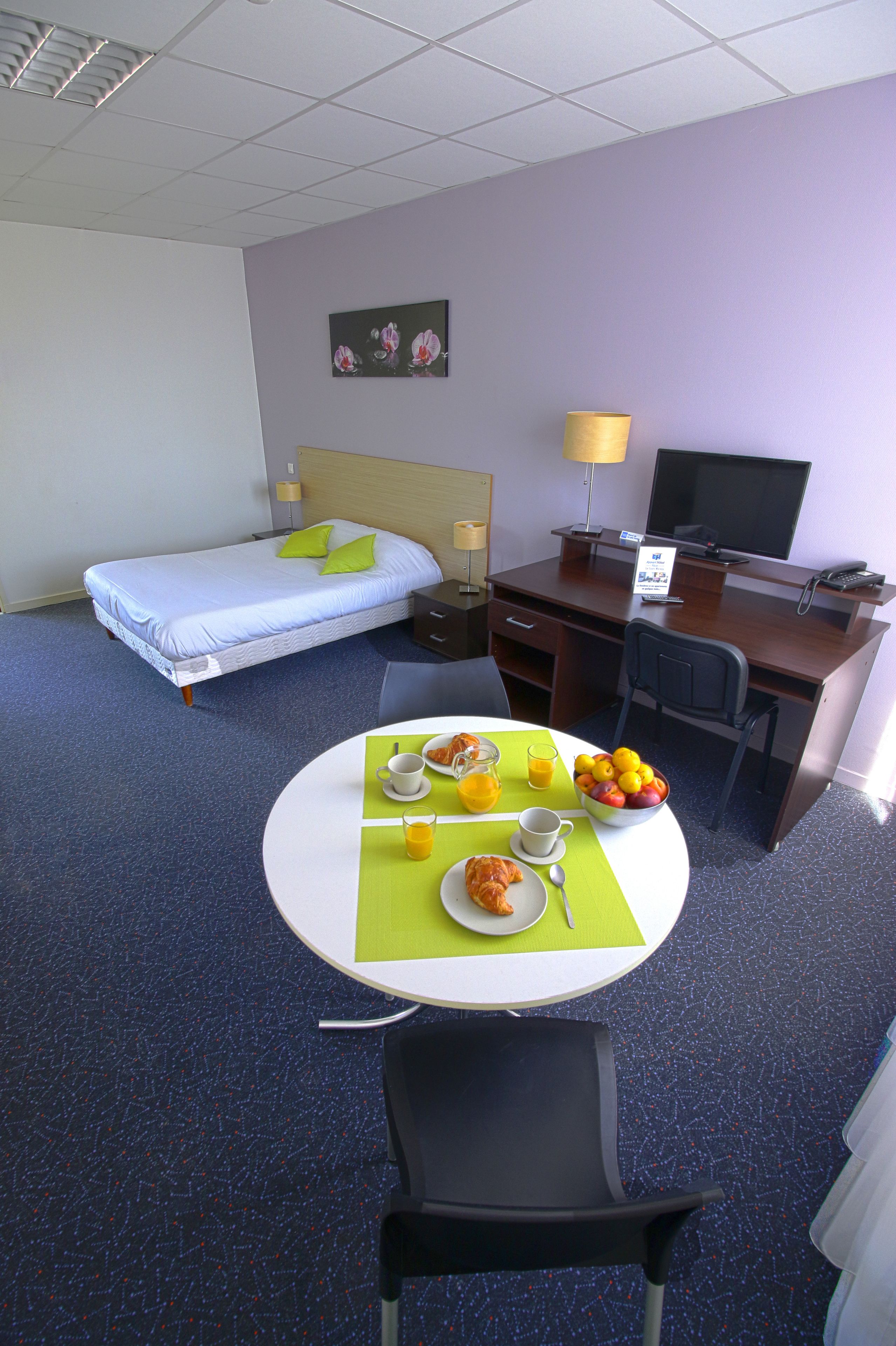 A hotel room with a bed, a desk, a chair, a table with breakfast items, and a television.