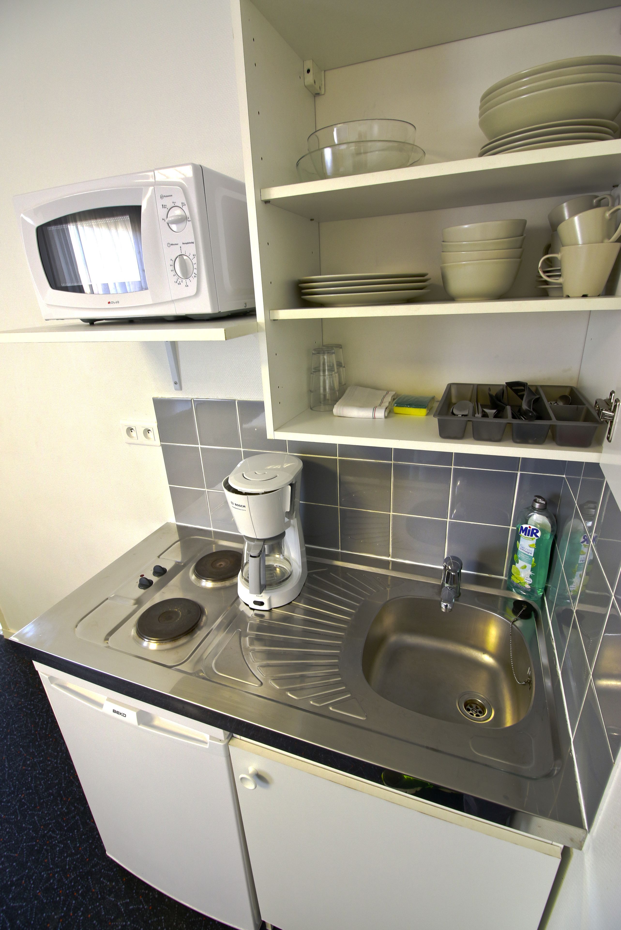 Een kleine keuken met een magnetron, koffiezetapparaat, spoelbak en servies.