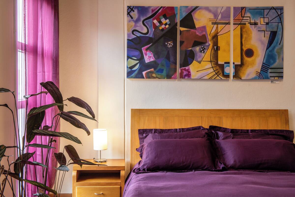 A hotel room with a bed, a nightstand, a lamp, a large abstract painting, and a potted plant.