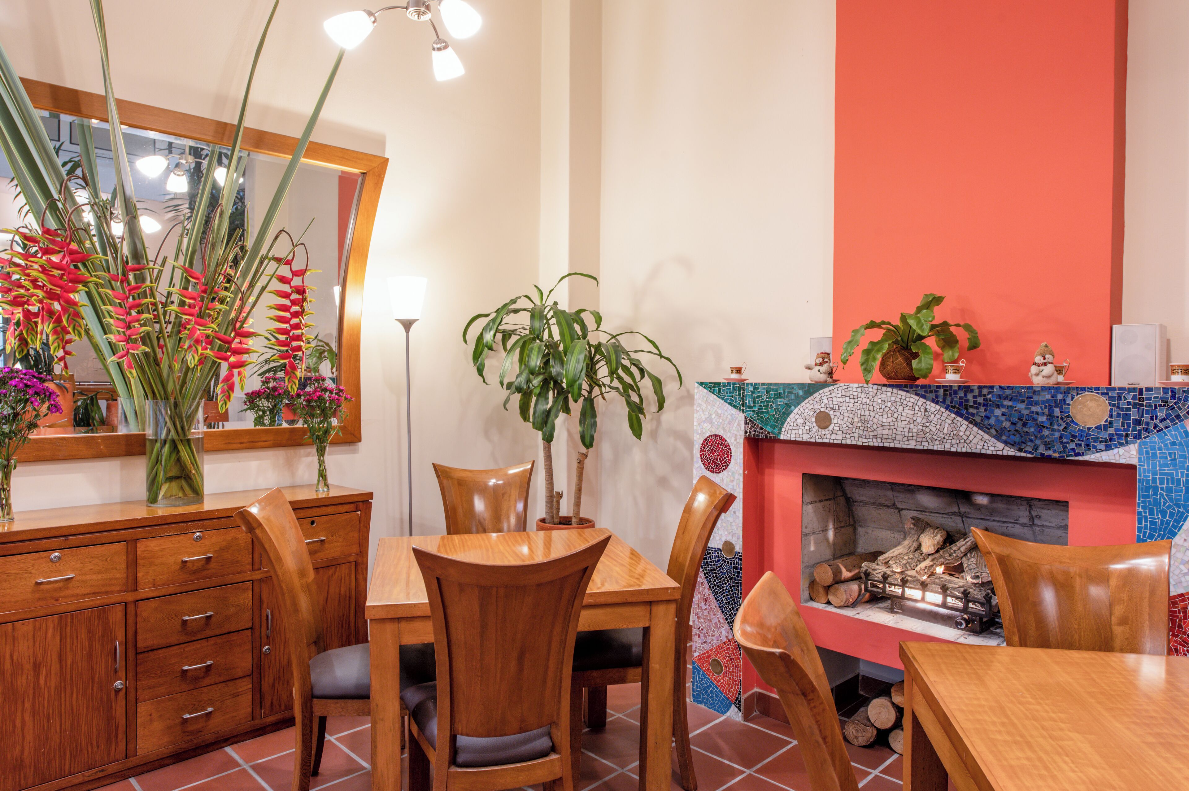 A dining area with a wooden table and chairs, a mirror, a potted plant, and a tiled backsplash with a fireplace.