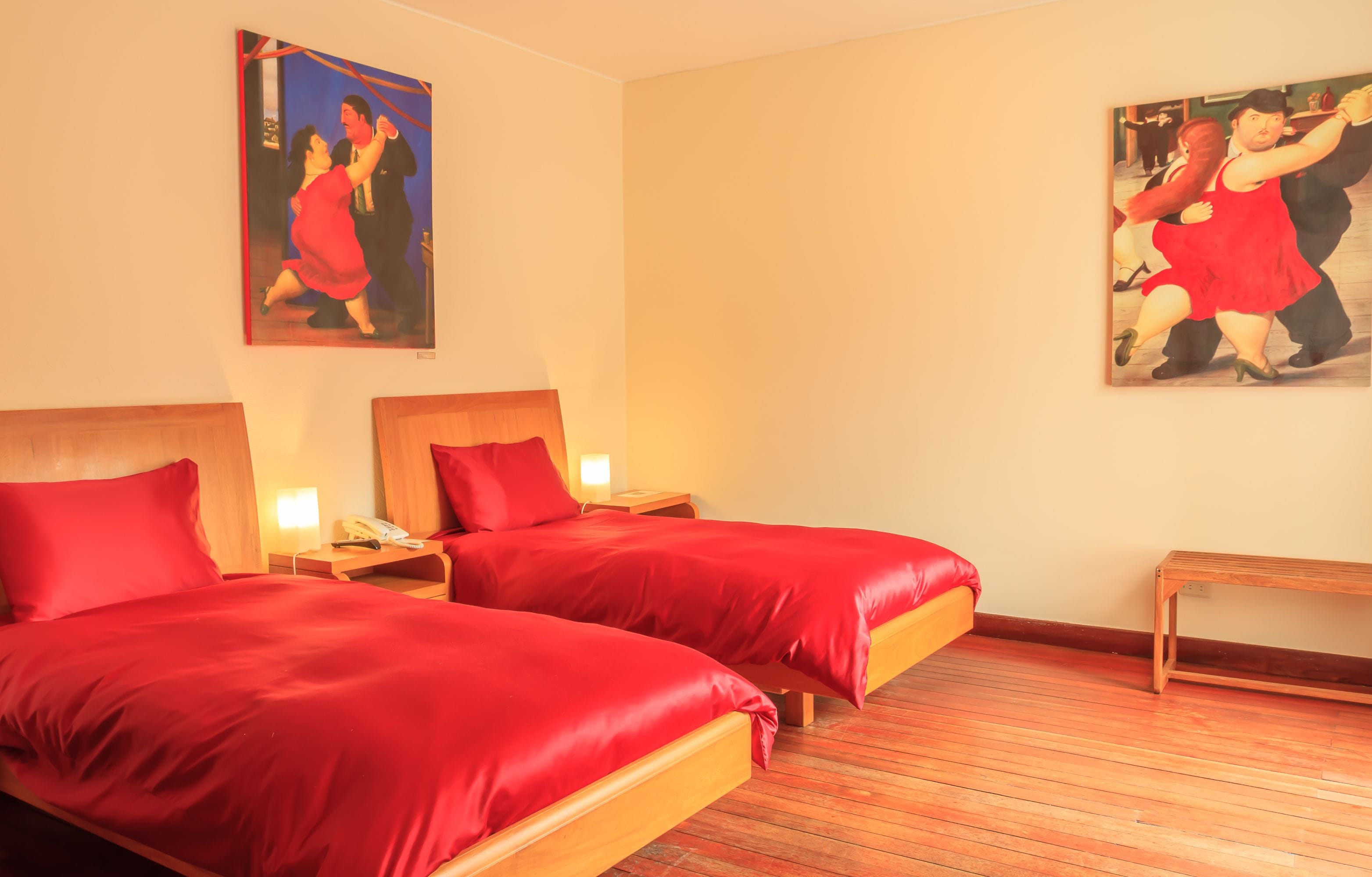 A hotel room with two beds, wooden headboards, red bedding, a wooden bench, and two paintings of dancing couples on the walls.