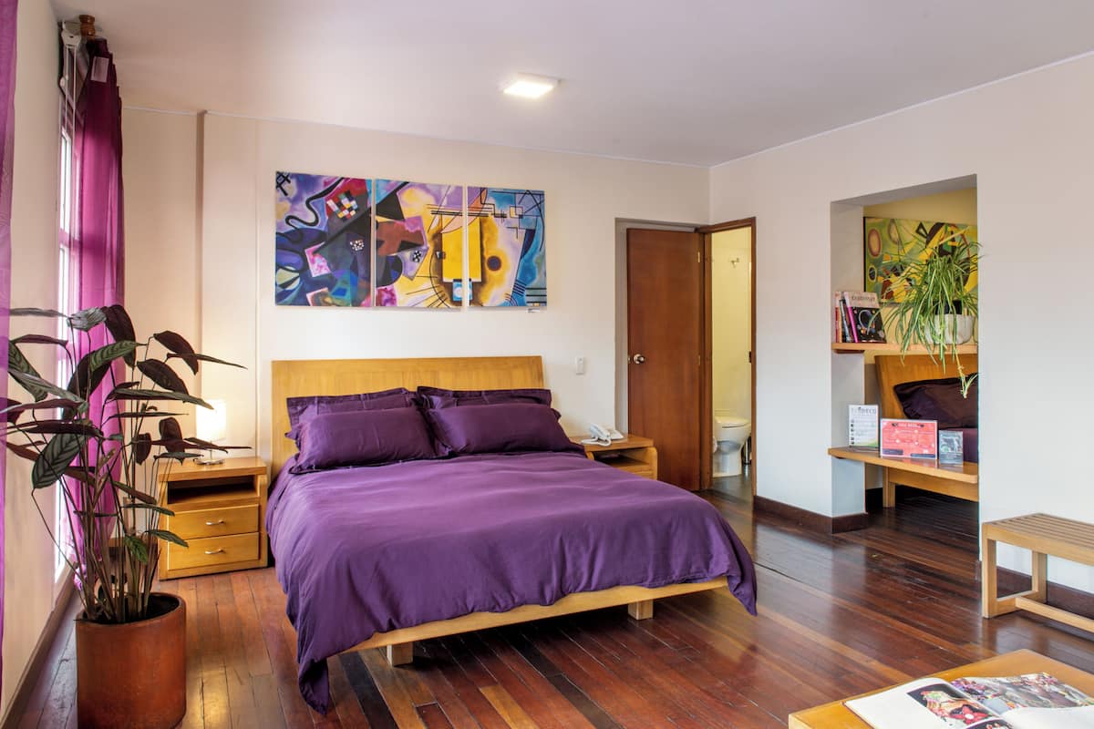 A bedroom with a wooden bed, purple bedding, a nightstand, a bedside table, a potted plant, and a wall with abstract art.