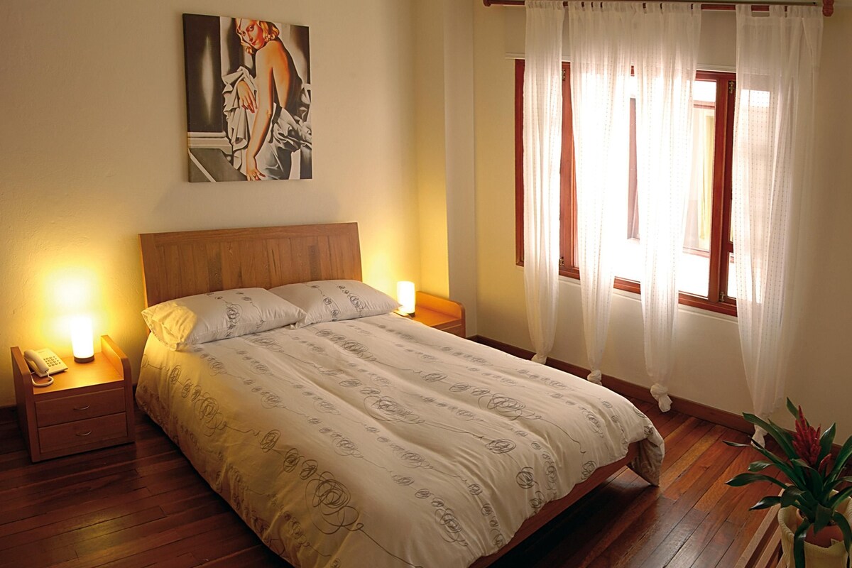 Un dormitorio con cama de madera, lámparas de noche, un cuadro en la pared y una ventana con cortinas.