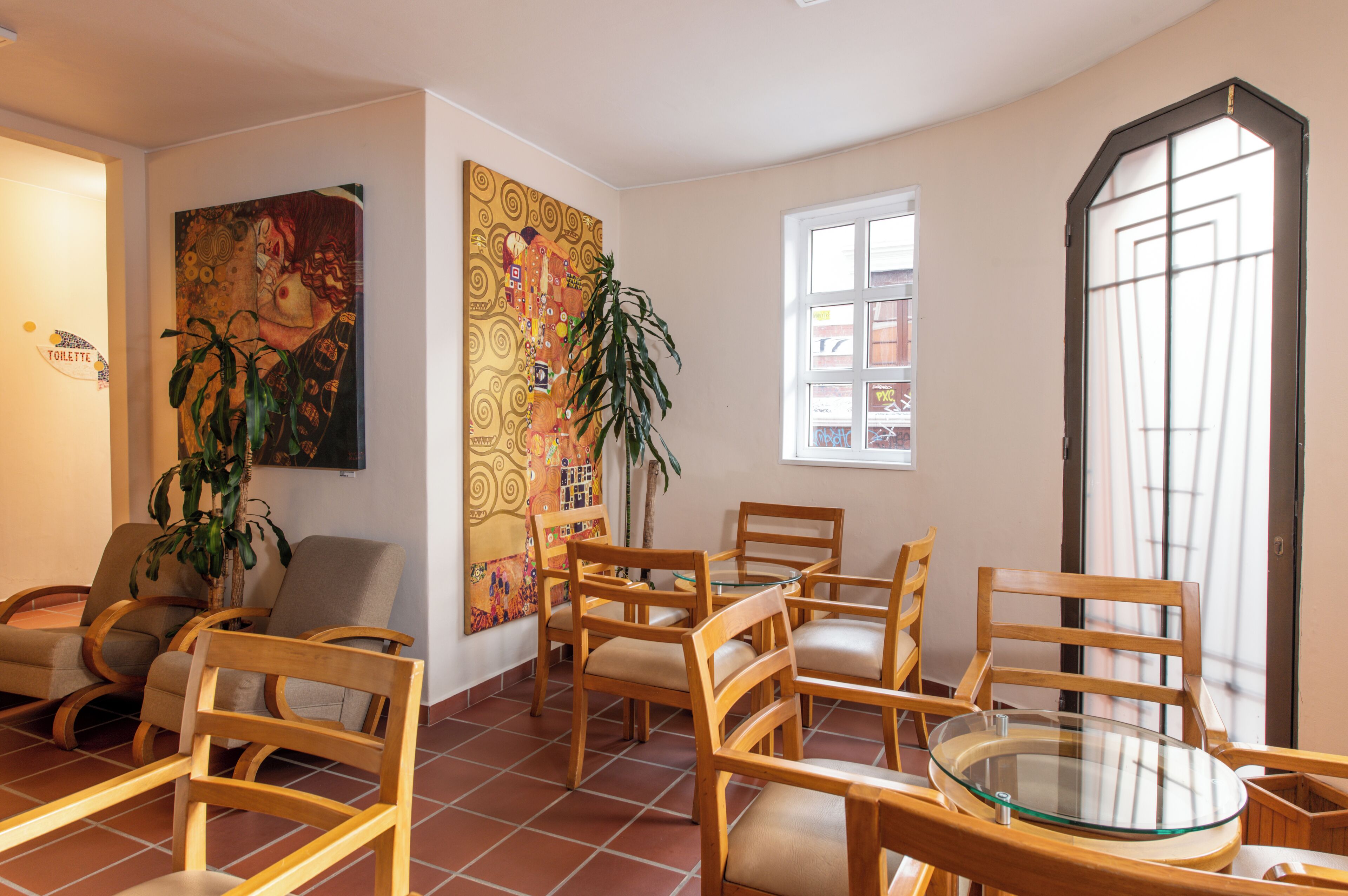 A room with wooden chairs and a glass table, a large painting on the wall, and a window with a view.