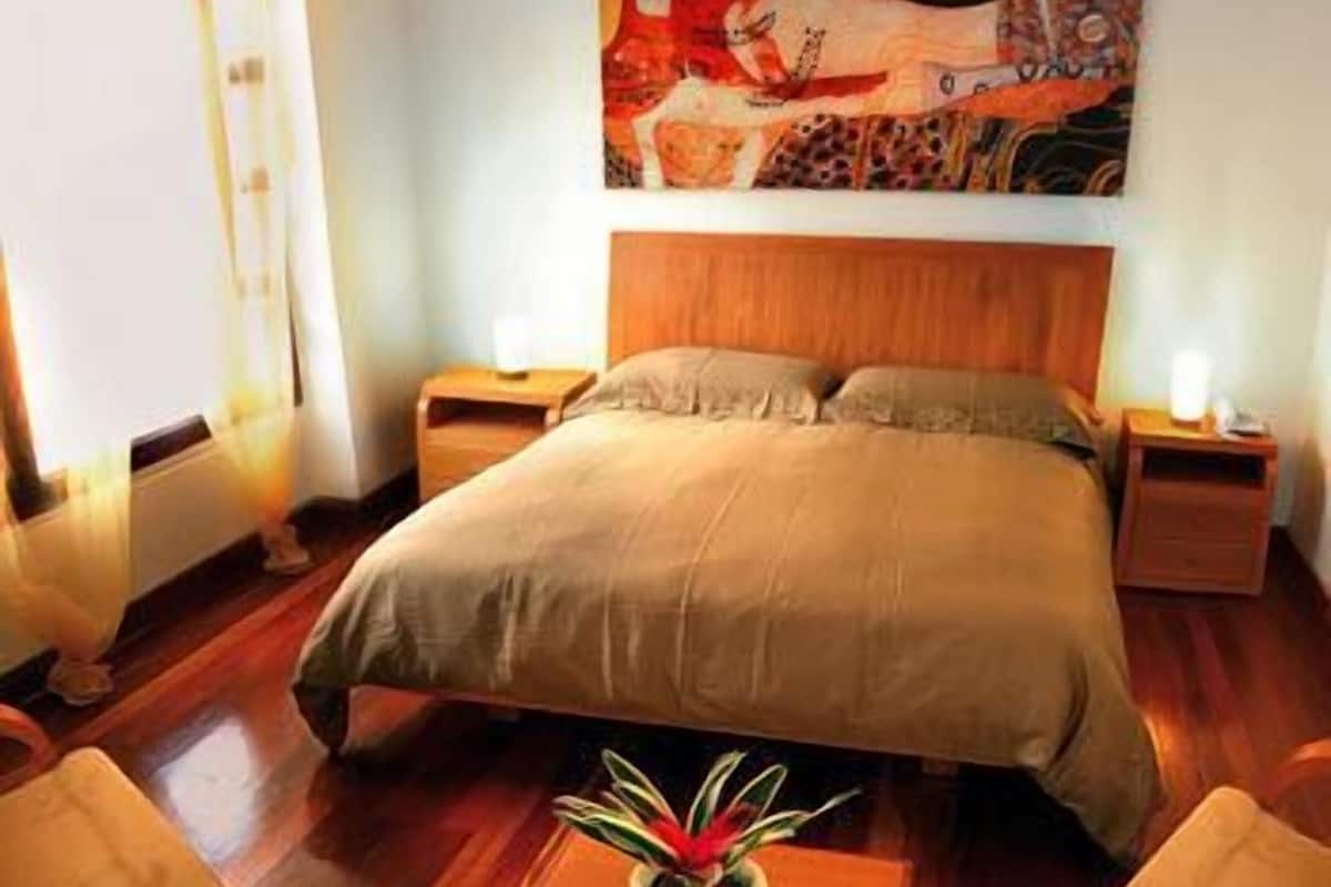 A bedroom with a large bed, two bedside tables, a nightstand, a potted plant, and a painting on the wall.