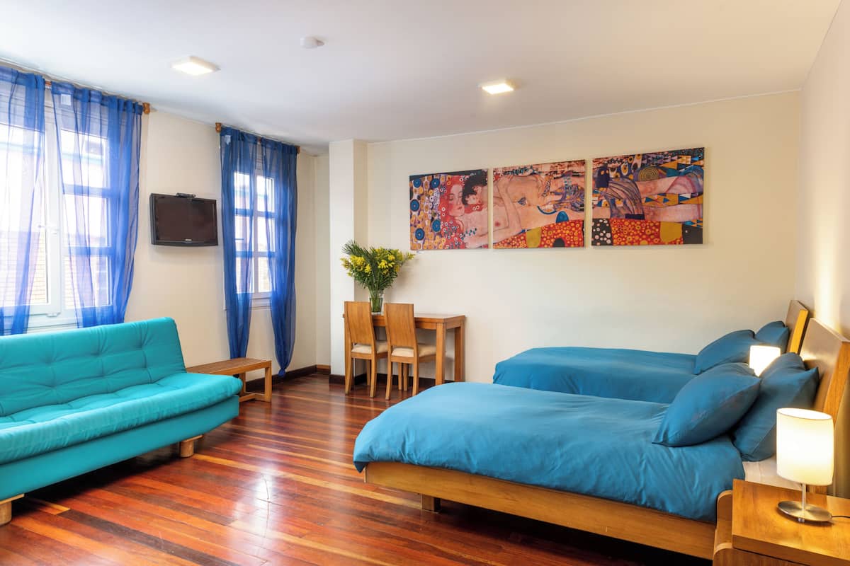 A modern hotel room with a wooden floor, a sofa, a bed with blue bedding, a desk with chairs, and two paintings on the wall.