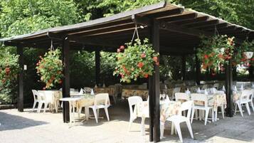 Eine Terrasse im Freien mit weißen Plastikstühlen und -tischen, überdacht von einer Holzpergola, umgeben von Grünpflanzen.