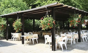 Une terrasse extérieure avec des chaises et des tables en plastique blanc, recouverte d’une pergola en bois et entourée de verdure.