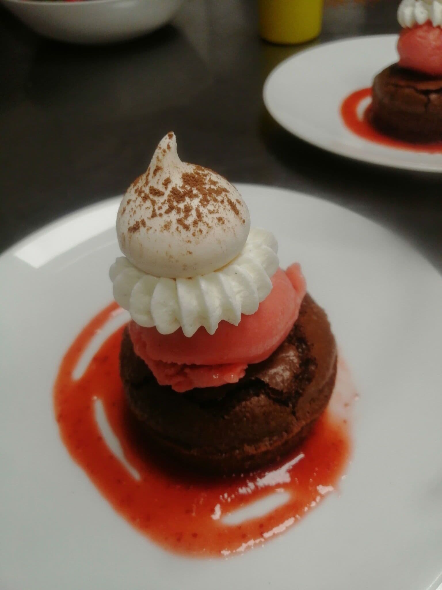 A dessert on a white plate with a chocolate base, topped with whipped cream and a scoop of pink ice cream, and drizzled with red sauce.