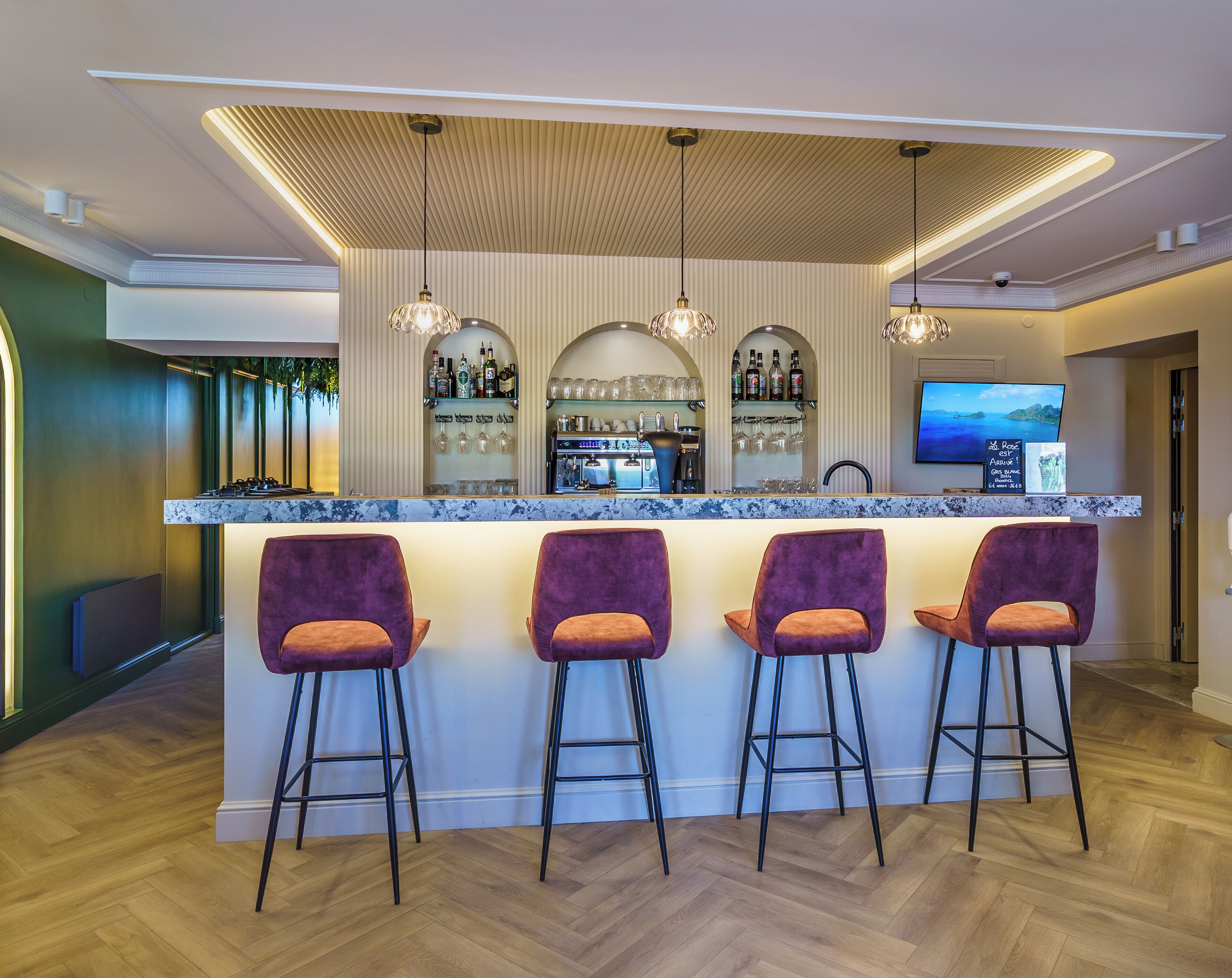 Una zona de bar con mostrador de mármol, tres taburetes morados, una barra con nevera para vino y un televisor montado en la pared.