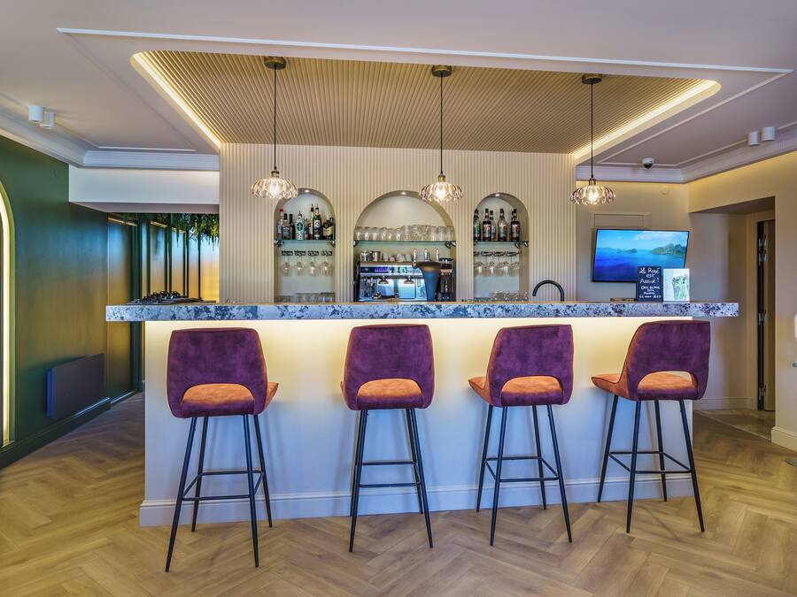 Una zona de bar con mostrador de mármol, tres taburetes morados, una barra con nevera para vino y un televisor montado en la pared.