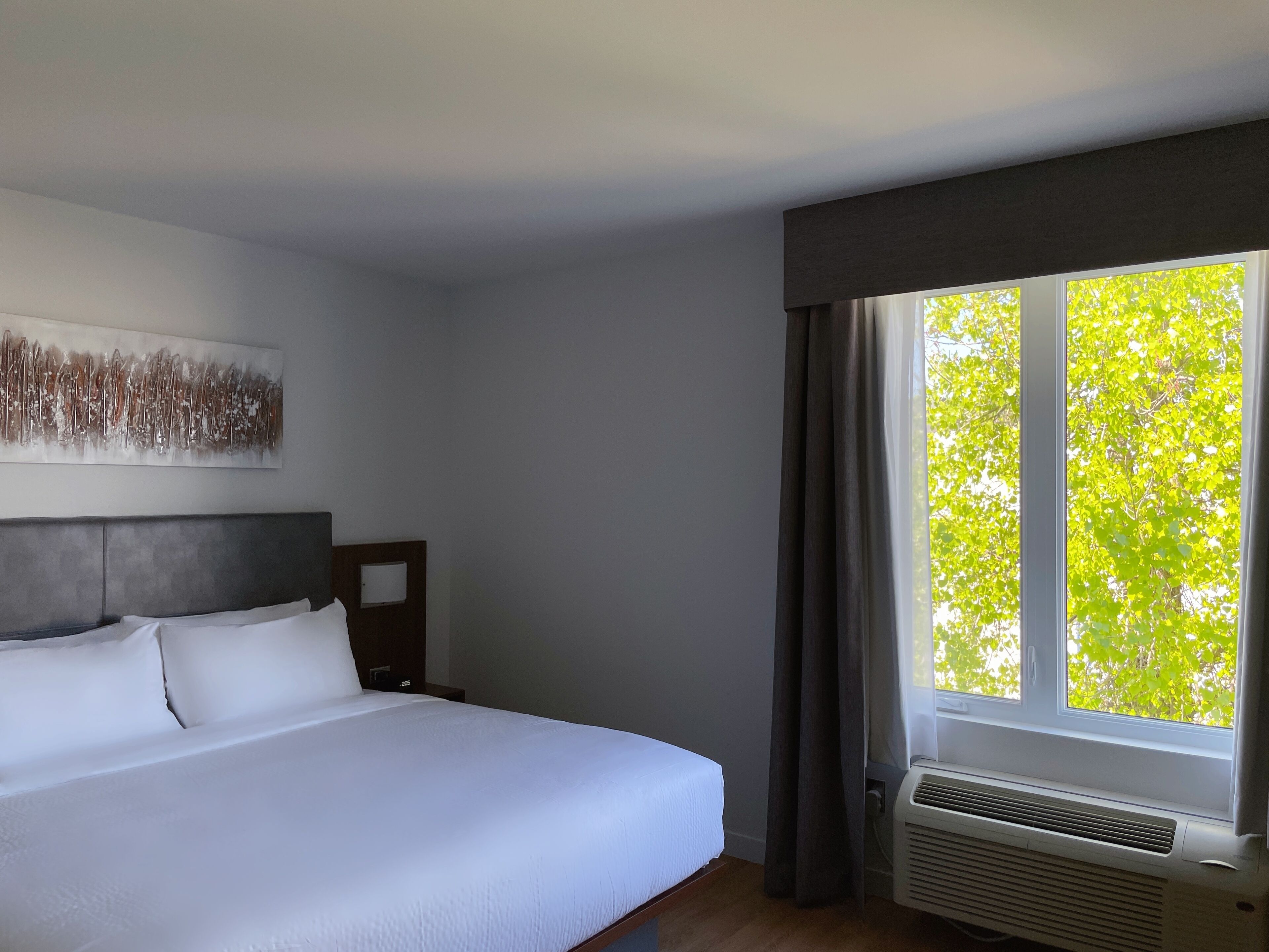 A hotel room with a large bed, a wall-mounted artwork, a window with curtains, and a built-in air conditioning unit.