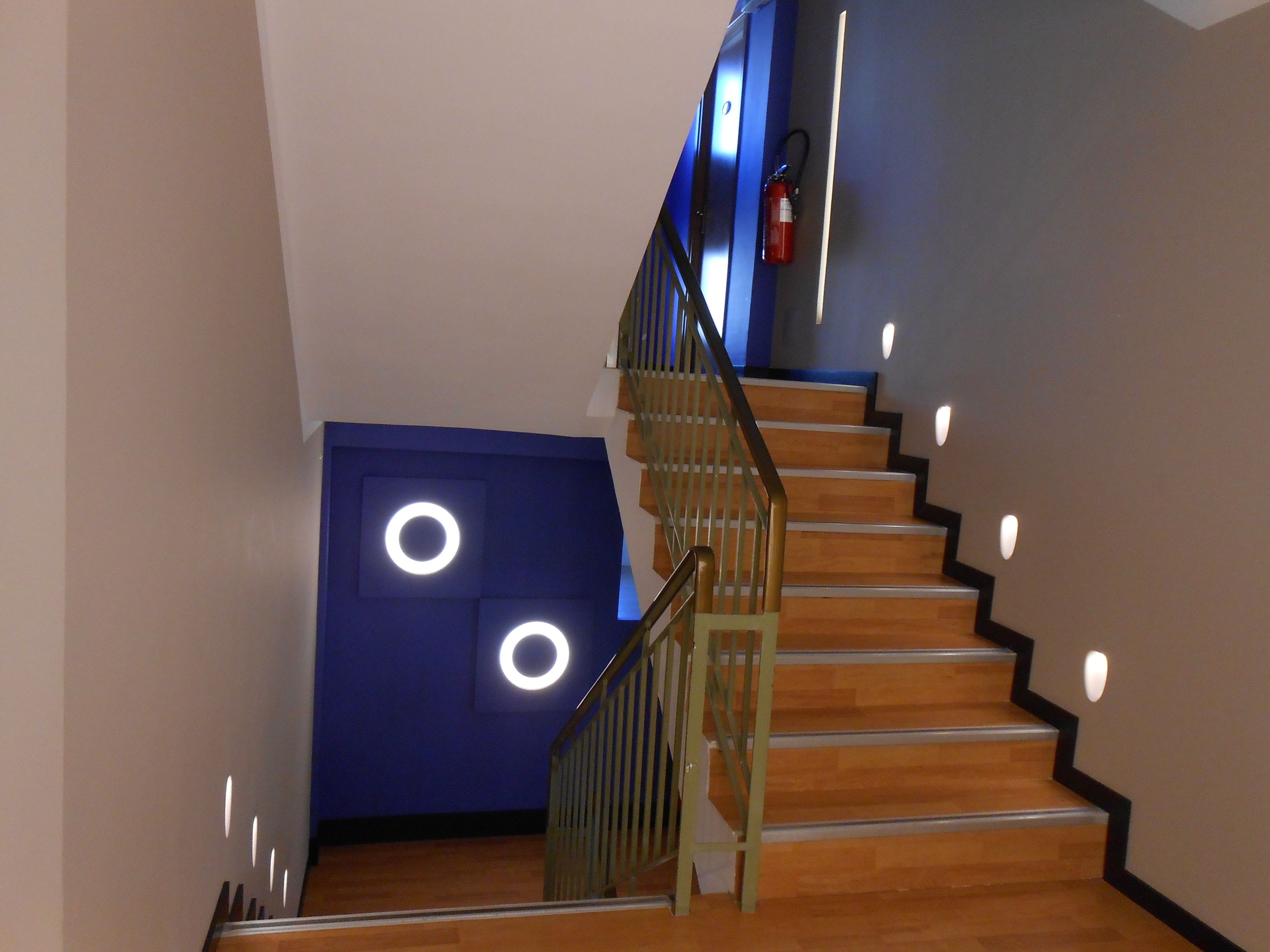 Un escalier avec des marches en bois et une rampe en métal, menant à un palier avec un mur bleu et des luminaires circulaires.