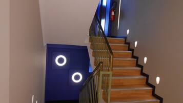 Un escalier avec des marches en bois et une rampe en métal, menant à un palier avec un mur bleu et des luminaires circulaires.