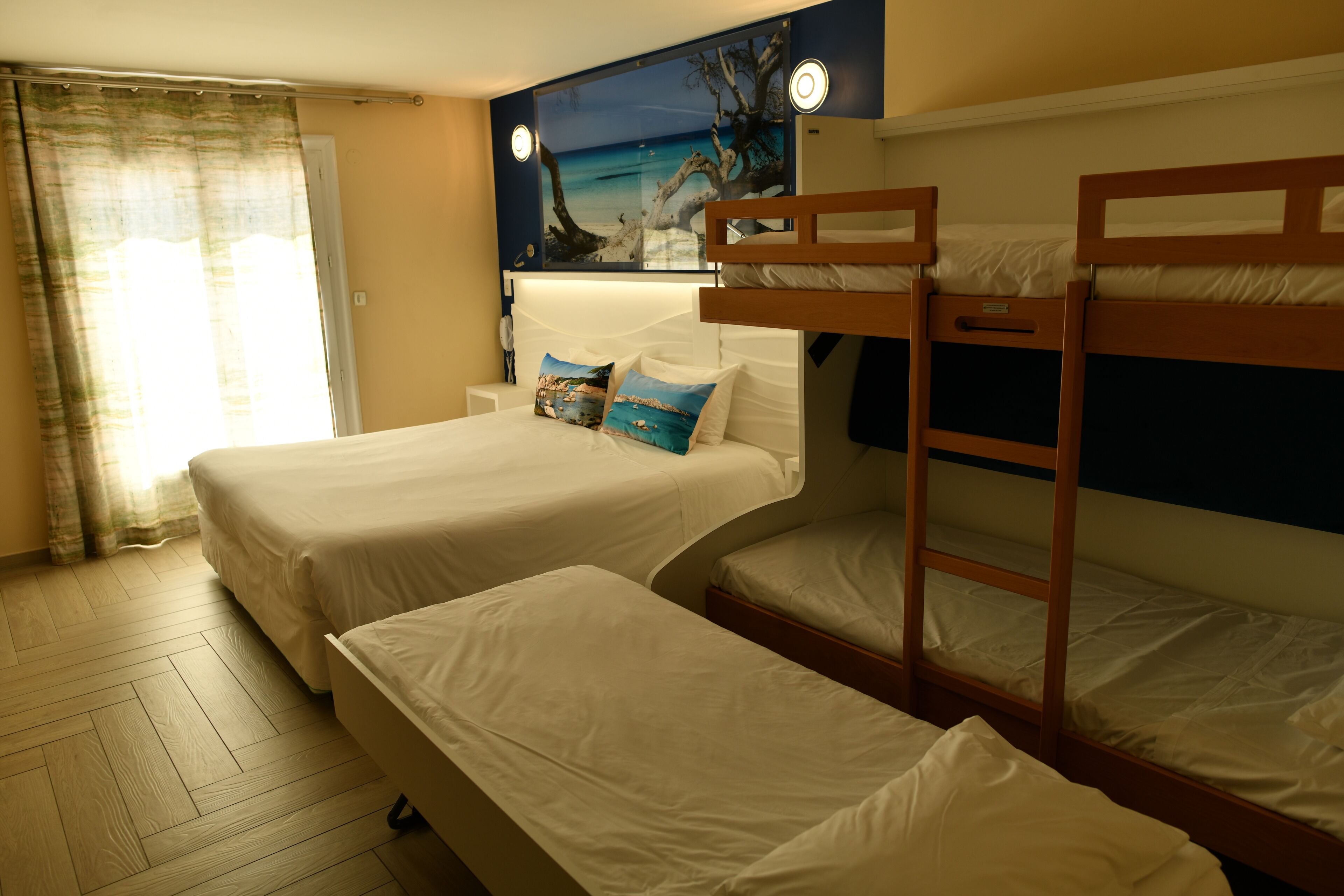 A hotel room with two single beds, a wooden bunk bed, a nightstand, a lamp, and a painting of a beach scene.