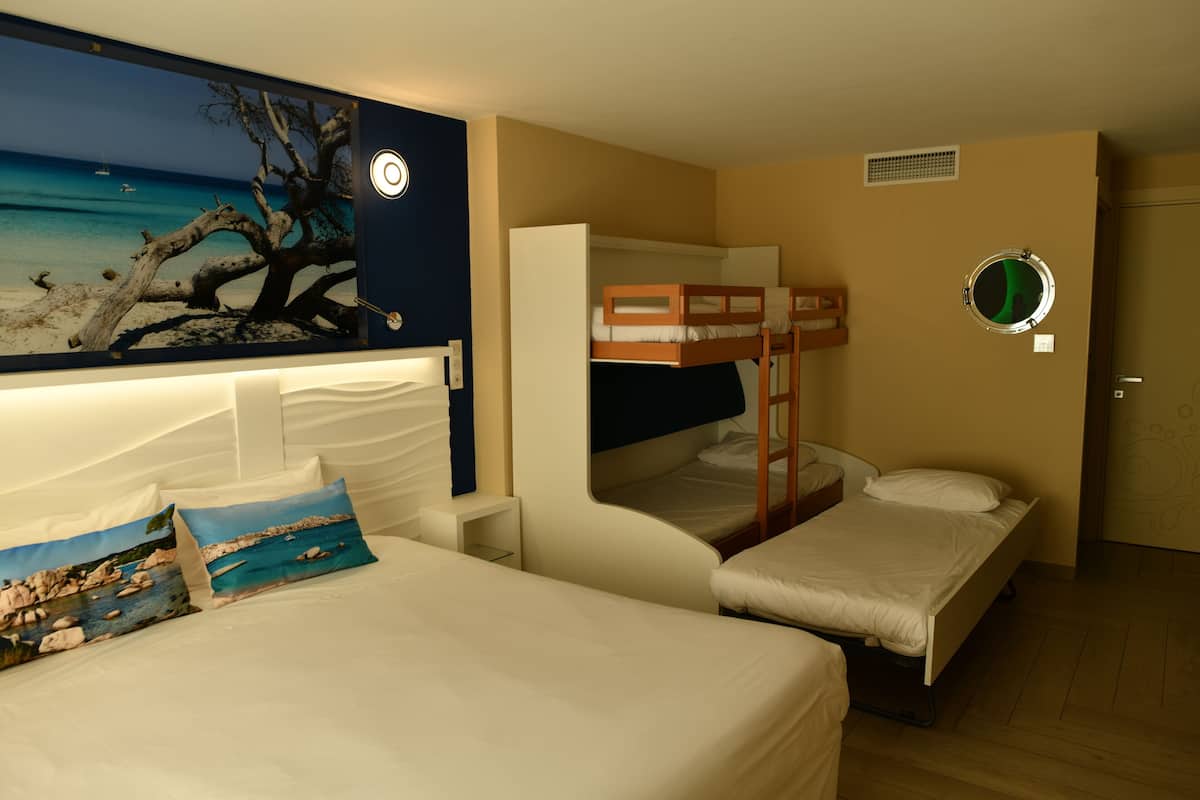 A room with two beds, one with a bunk bed and another single bed, a large window with a view of the sea, and a wall-mounted clock.